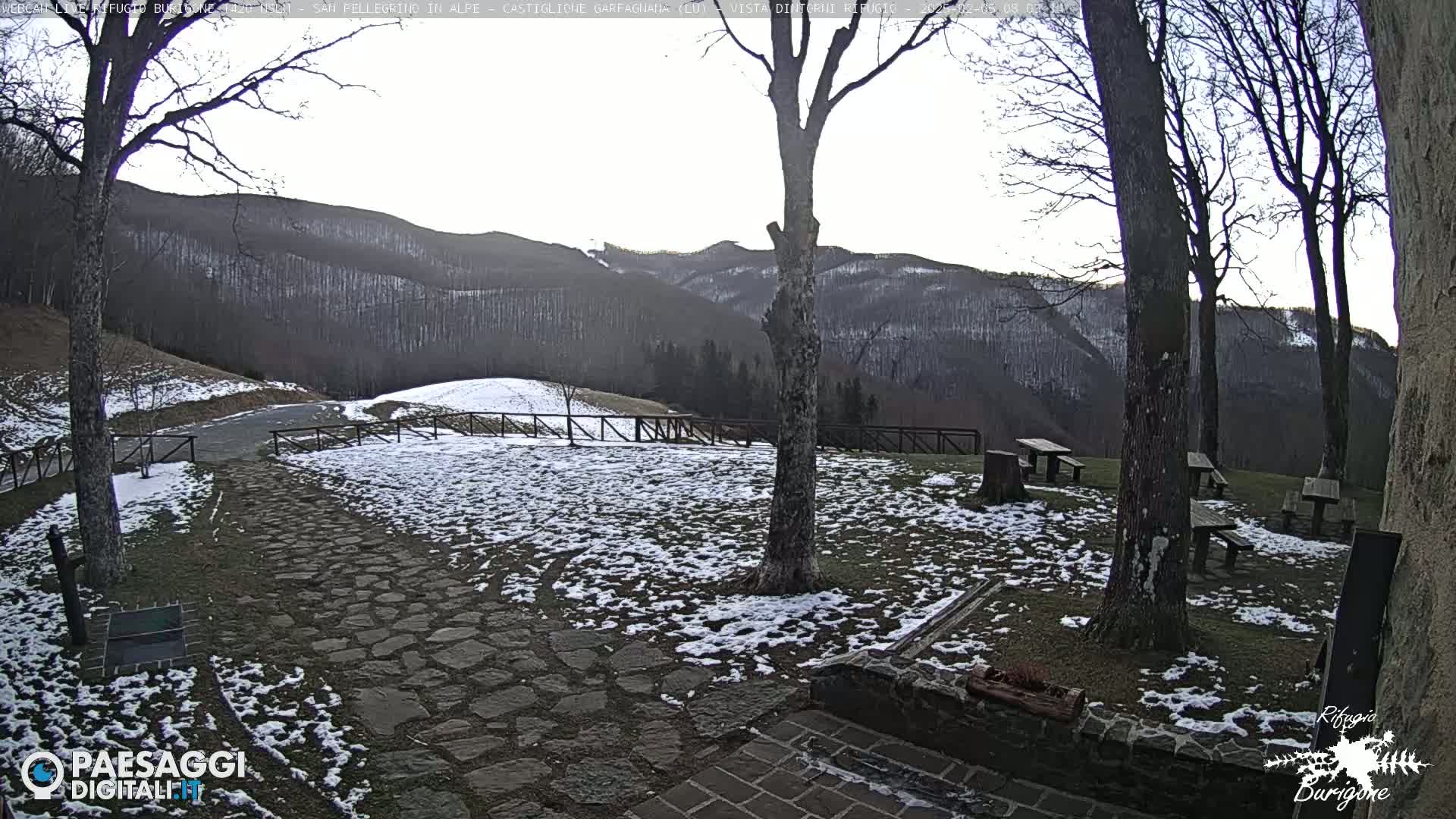 A stone pathway and picnic tables are partially covered in snow, with leafless trees and distant snow-covered mountains visible under a mostly clear sky.
