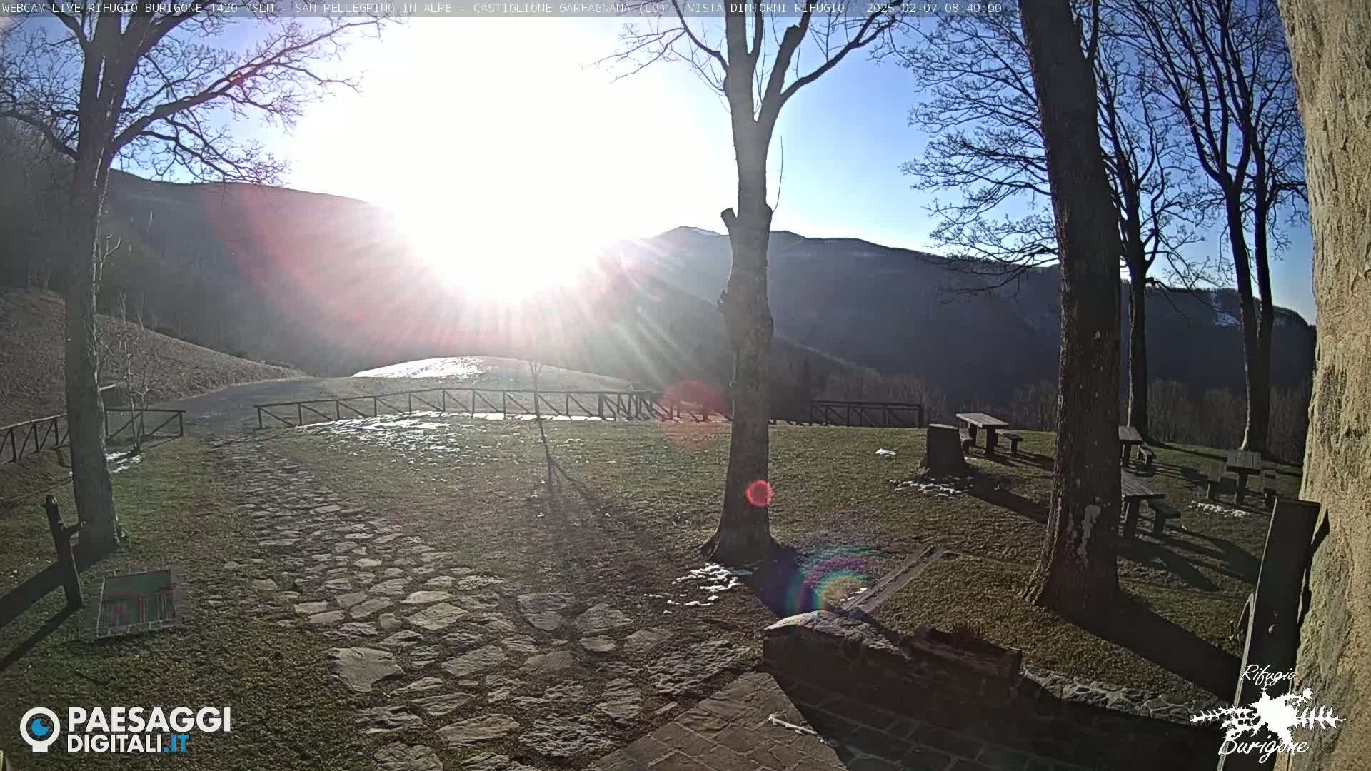 A sunny, cold day with patches of snow in a mountainous area shows a stone path leading to picnic tables near bare trees, with a bright sun shining over a distant hill.