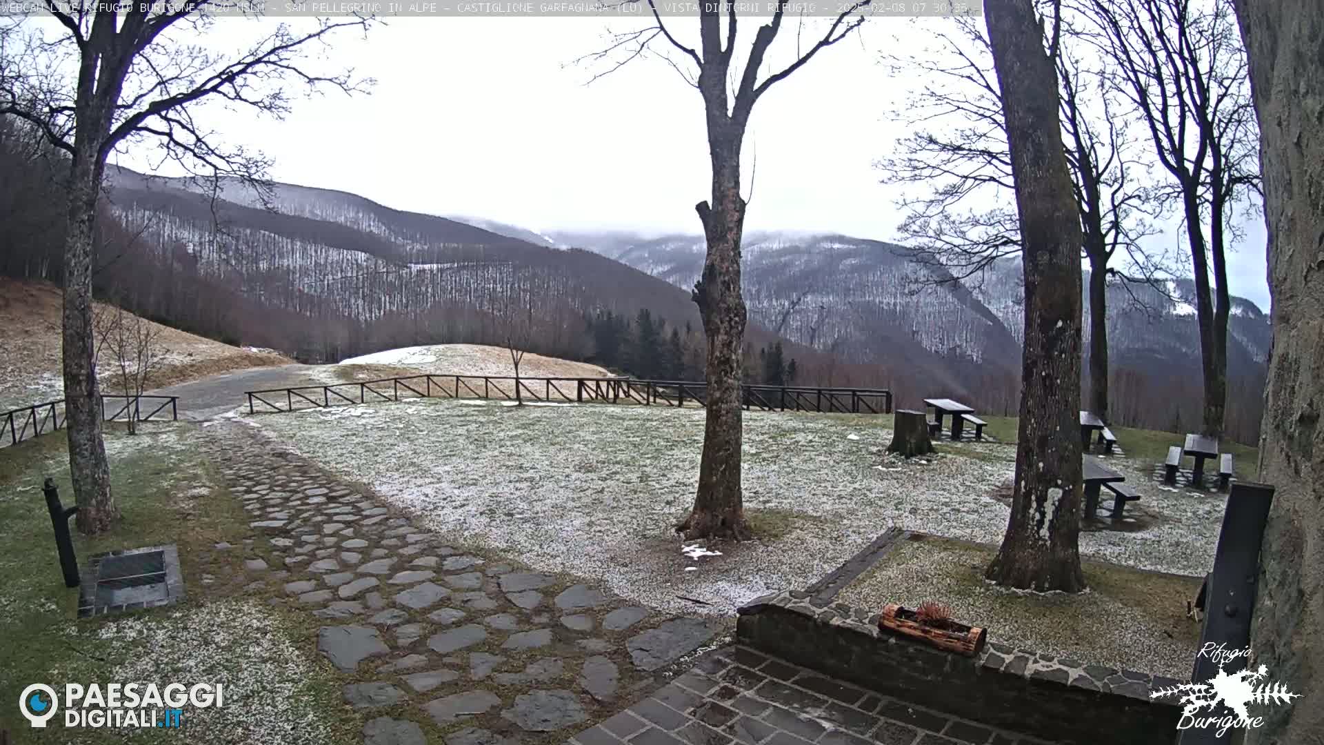 A snow-dusted stone path leads to picnic tables situated in a grassy area overlooking a mountain range under a cloudy sky.