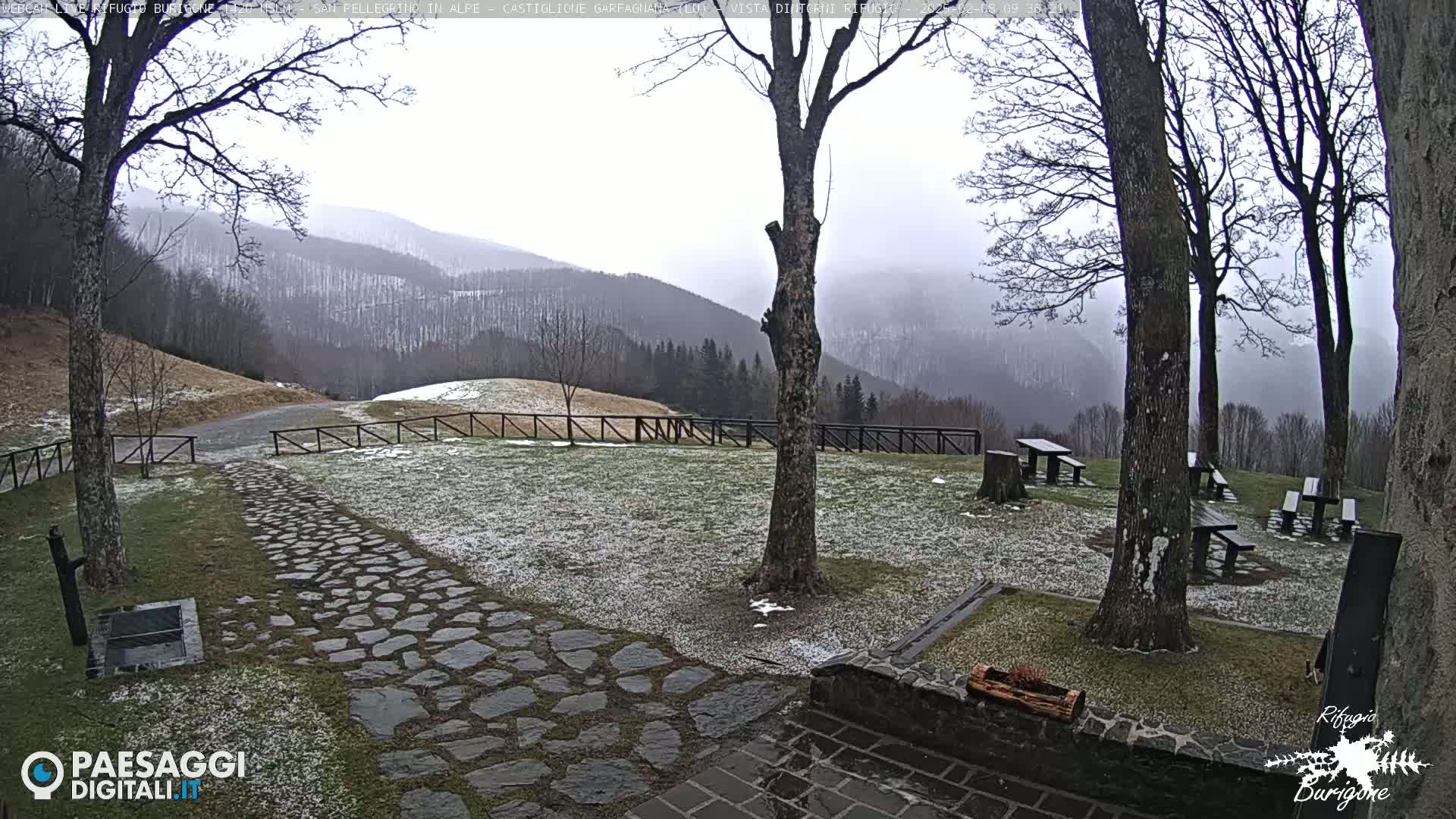 A snow-dusted stone path leads to several picnic tables situated in a grassy area overlooking a misty mountain range.