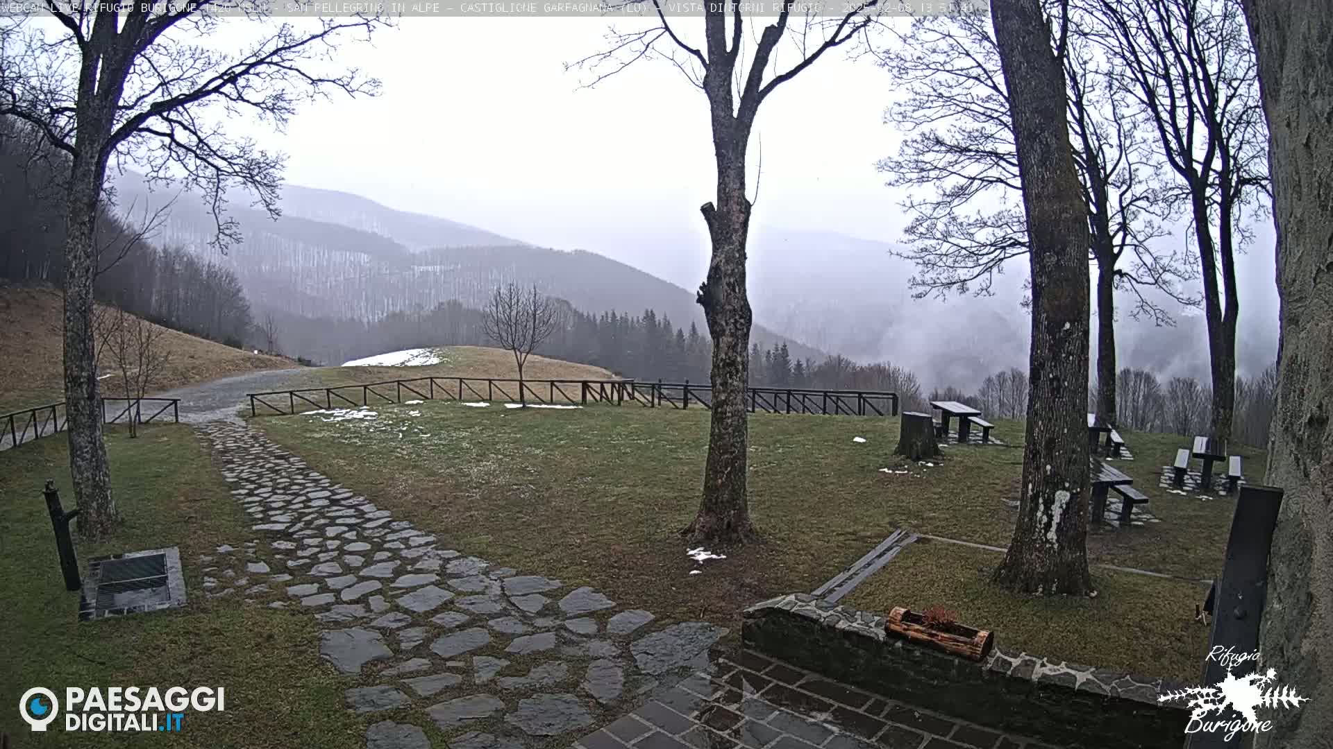A stone pathway leads to a grassy area with picnic tables under bare trees, overlooking a misty mountain range with patches of snow.