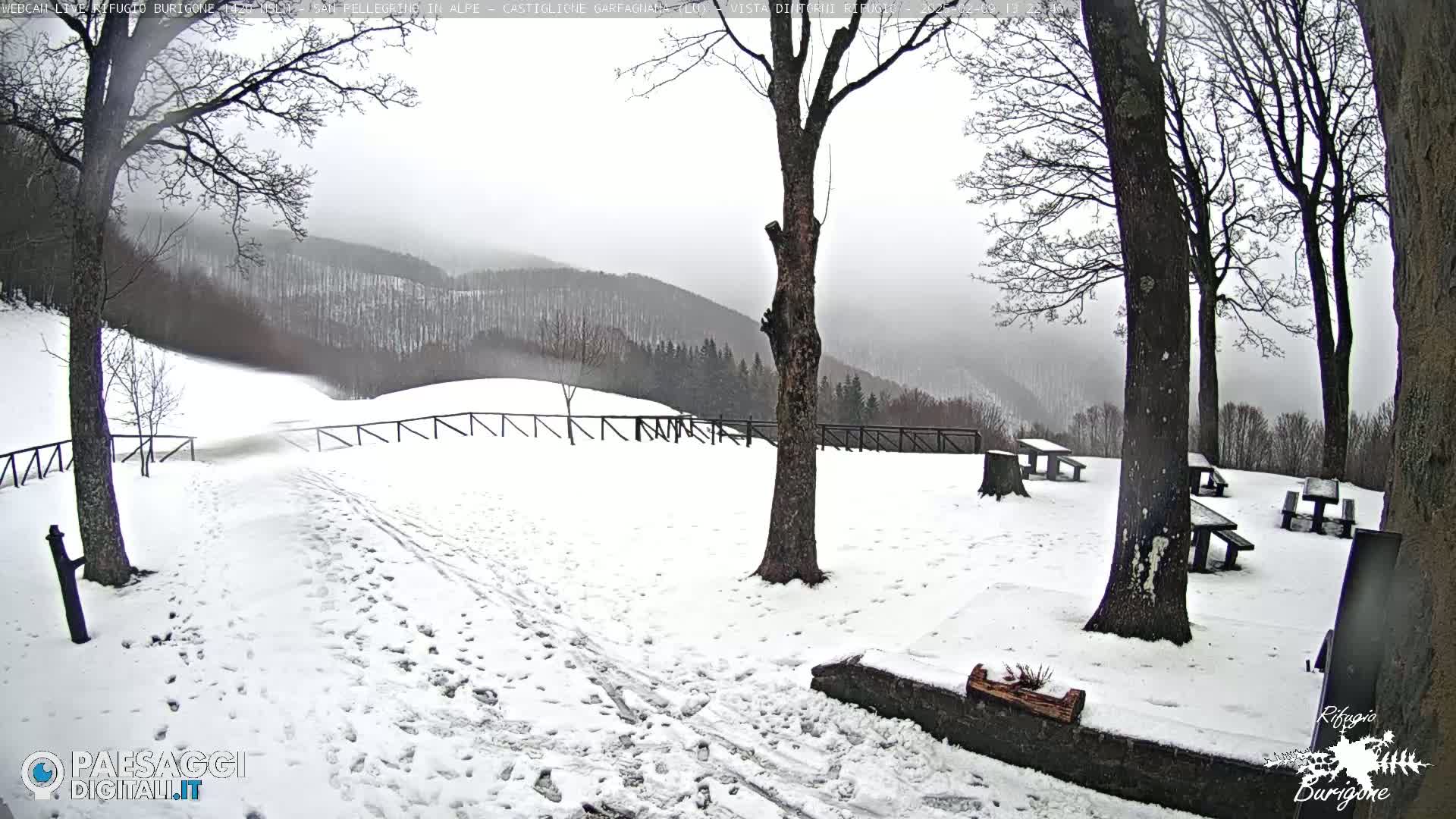 A snow-covered picnic area with several empty tables sits in front of a snow-dusted mountain range under a gray, overcast sky.