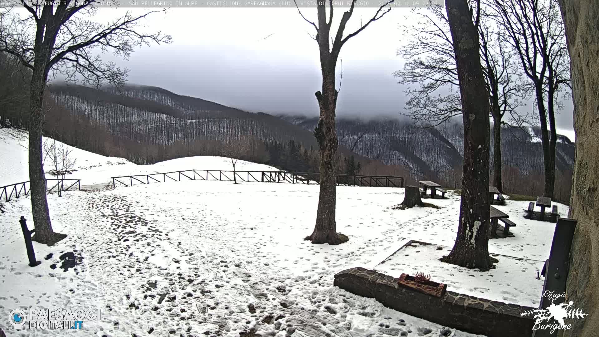 A snow-covered area with picnic tables and trees, overlooking a mountain range under a cloudy sky.