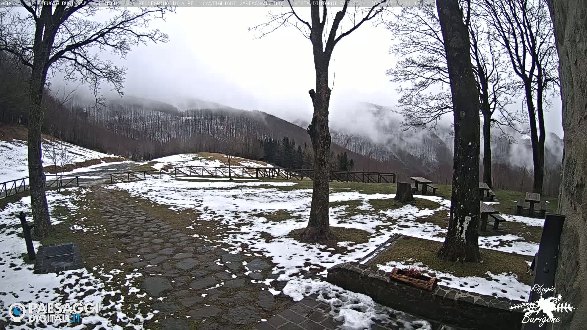A snow-dusted stone pathway leads to picnic tables situated beneath bare trees overlooking a misty mountain range.