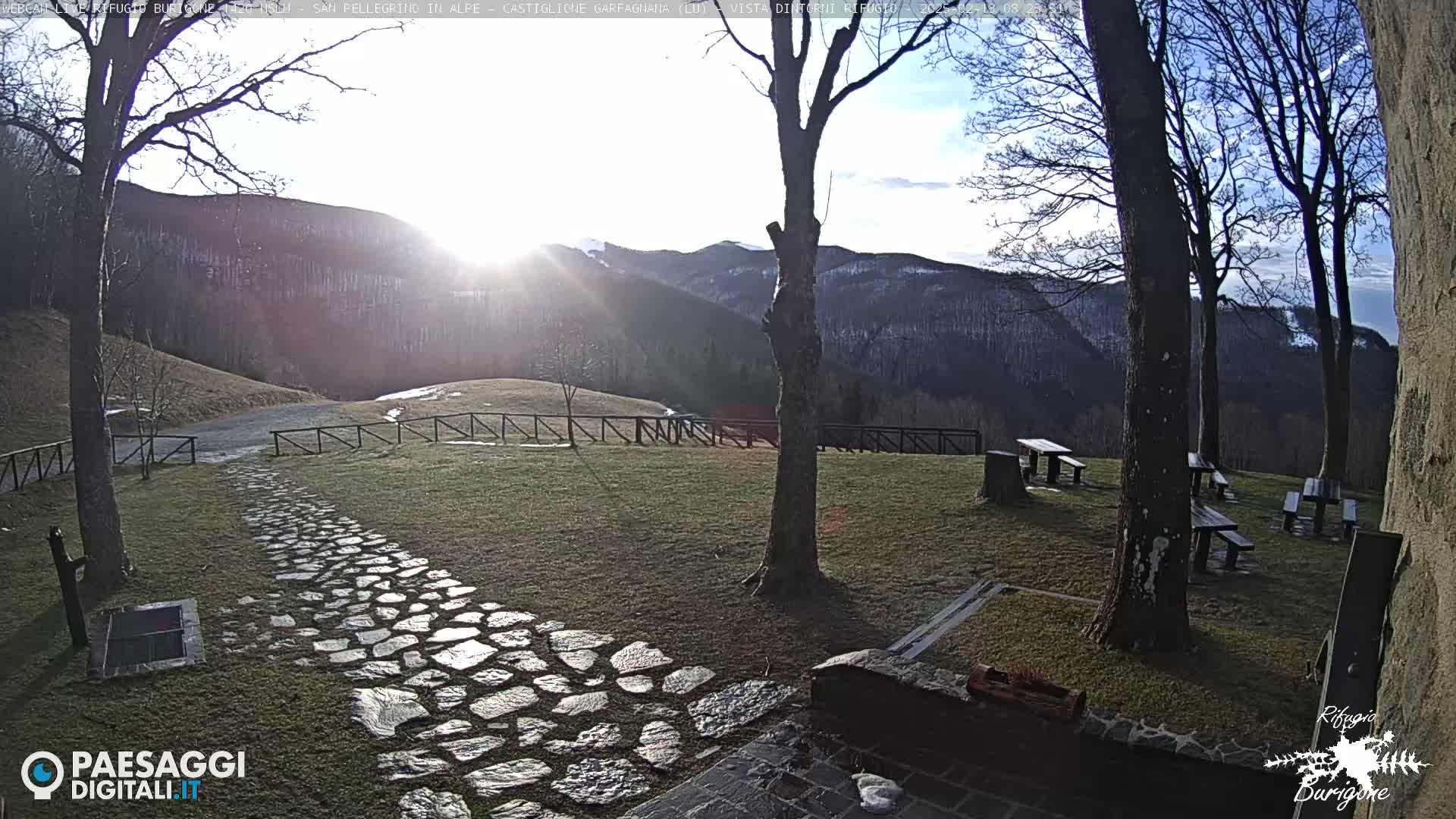 A stone path leads to several picnic tables situated on a grassy area overlooking a sunlit mountain range under a mostly clear sky.