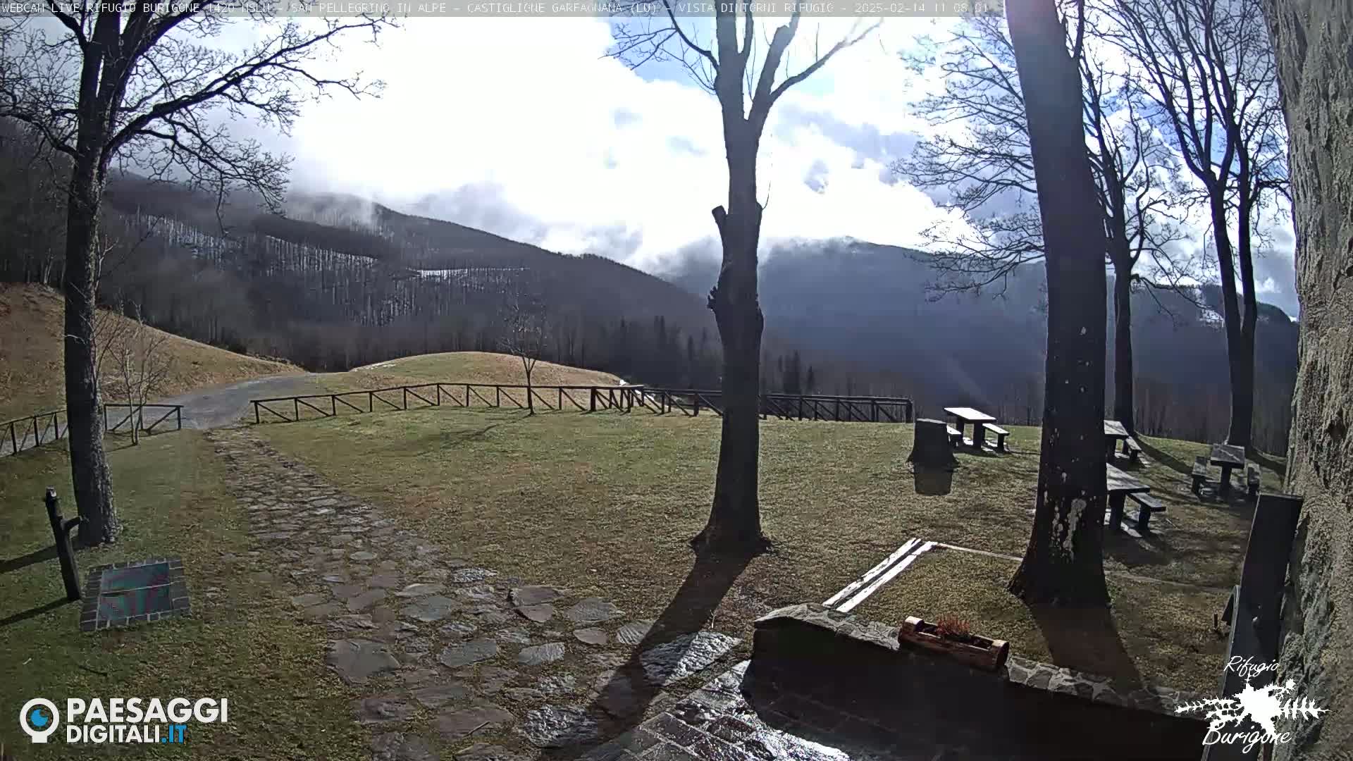 A grassy area with picnic tables and a stone path leading to a road, overlooks a mountain range partially covered in clouds and snow under a partly sunny sky.
