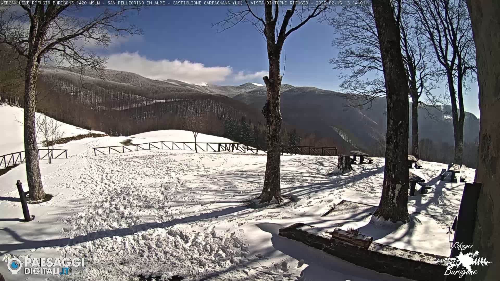 A snow-covered landscape with several picnic tables and trees under a mostly sunny sky, mountains in the background.