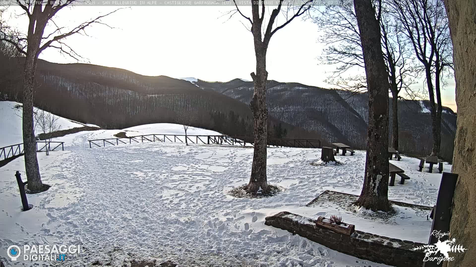 A snow-covered picnic area with several wooden tables and benches sits beneath leafless trees, overlooking a hilly landscape under a mostly clear sky.