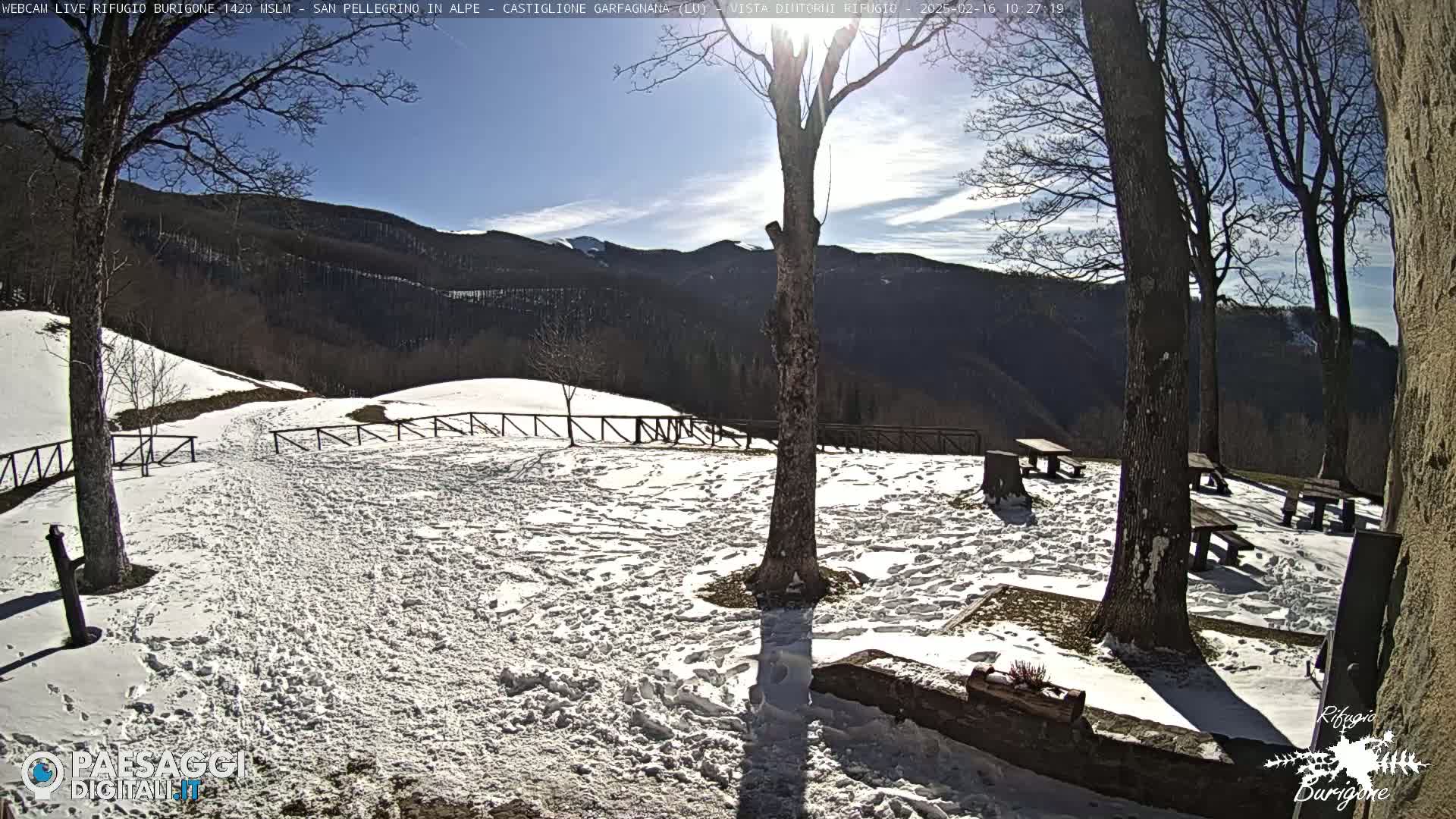 A snow-covered picnic area with several empty picnic tables is situated in front of a mountain range under a mostly sunny sky.