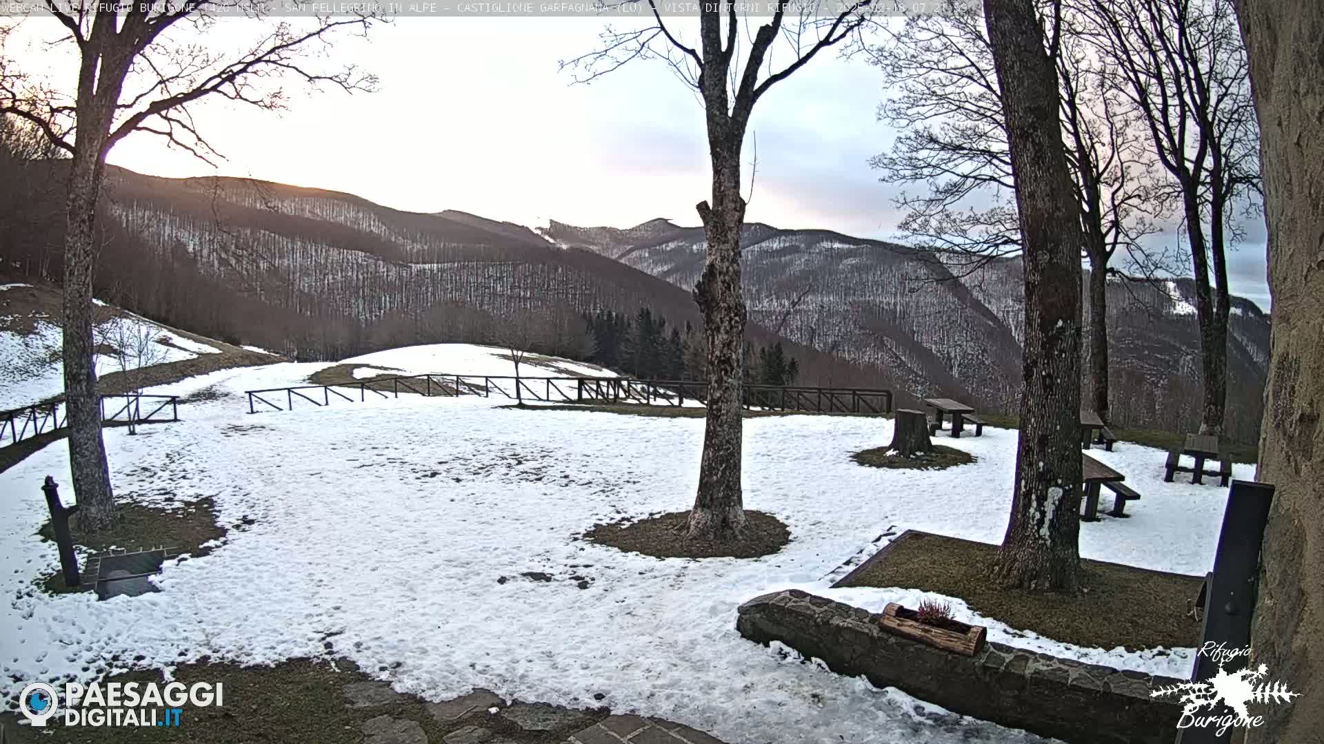 A snow-covered area with picnic tables and trees sits before a mountainous landscape under a cloudy sky.