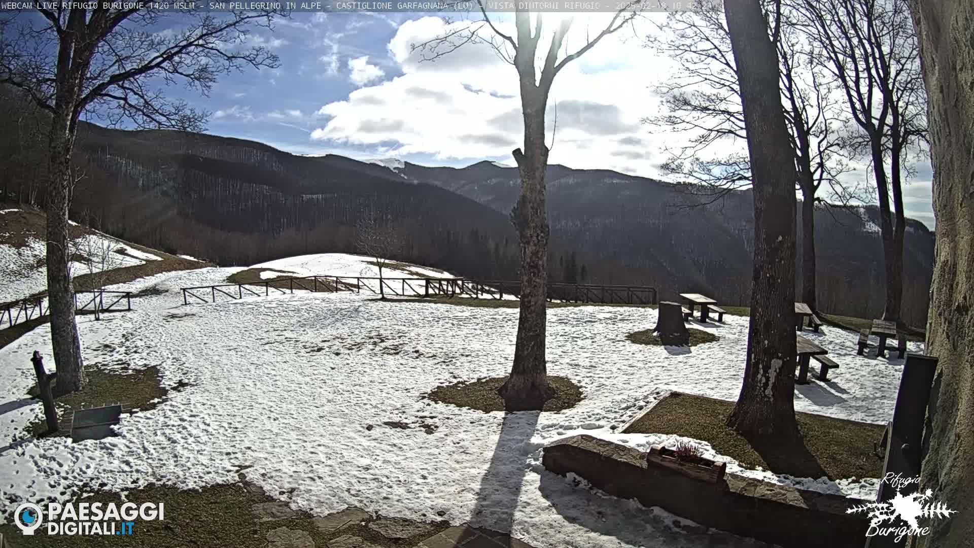 A snow-covered outdoor area with picnic tables, surrounded by bare trees and mountains under a mostly sunny sky.