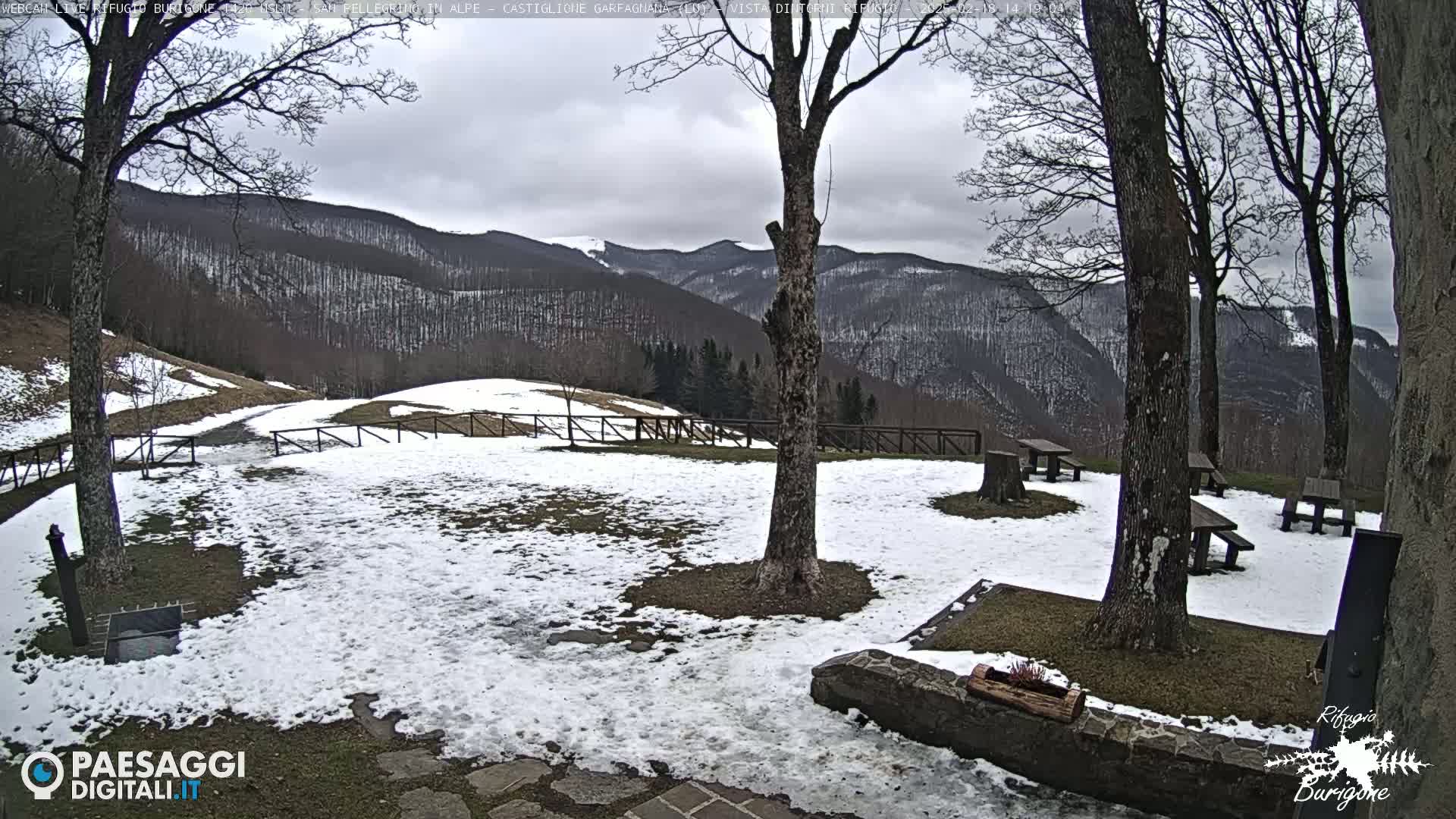 A snow-dusted outdoor area with picnic tables, trees, and a mountain range in the background under an overcast sky.
