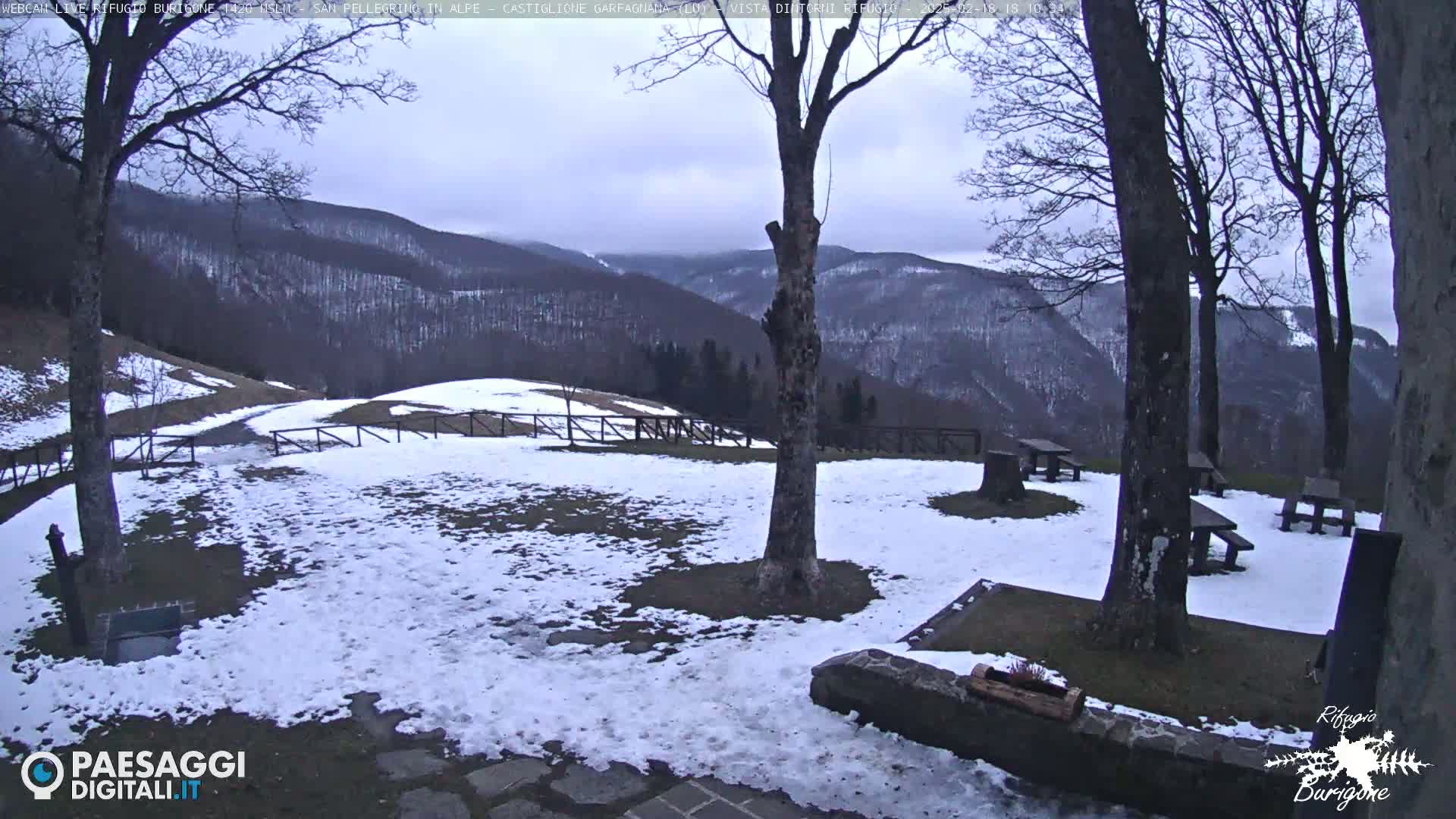 A snow-covered outdoor area with picnic tables and several bare trees overlooks a mountain range under an overcast sky.