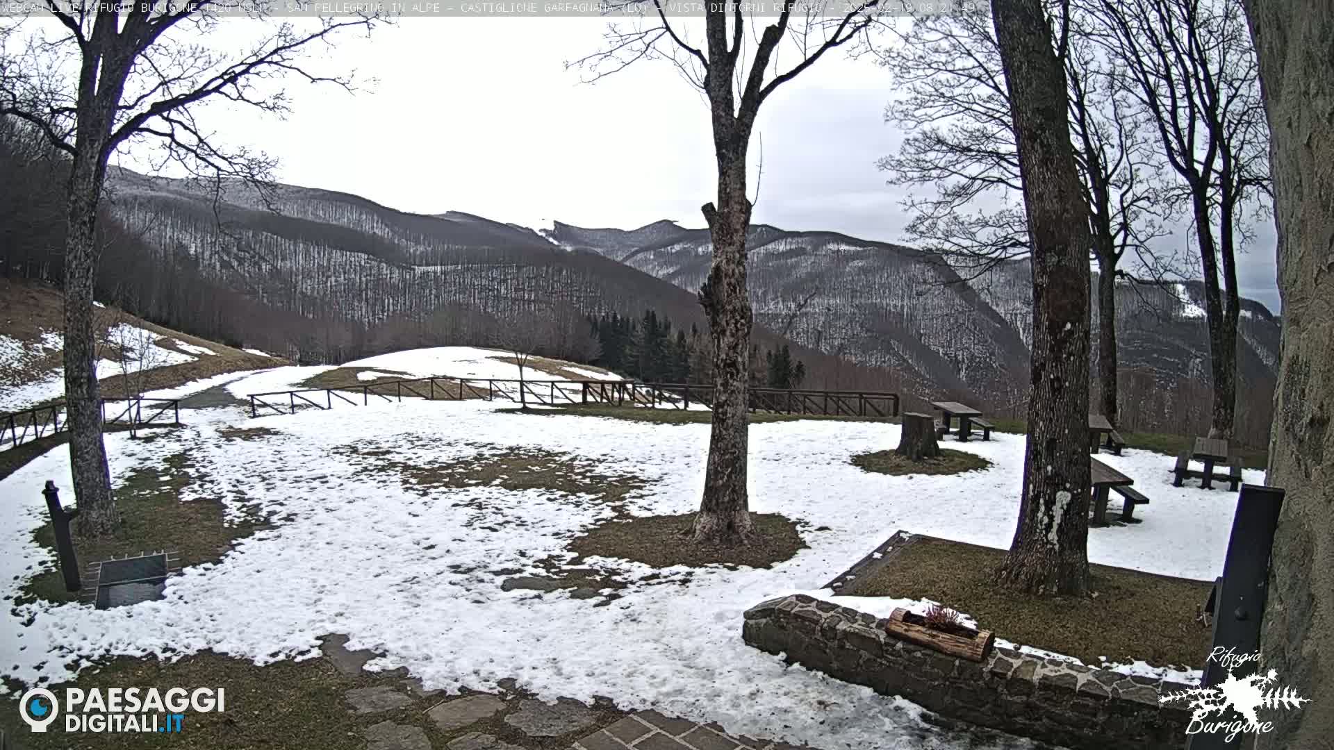 A snow-dusted outdoor area with picnic tables and several trees, overlooks a mountain range under a cloudy sky.