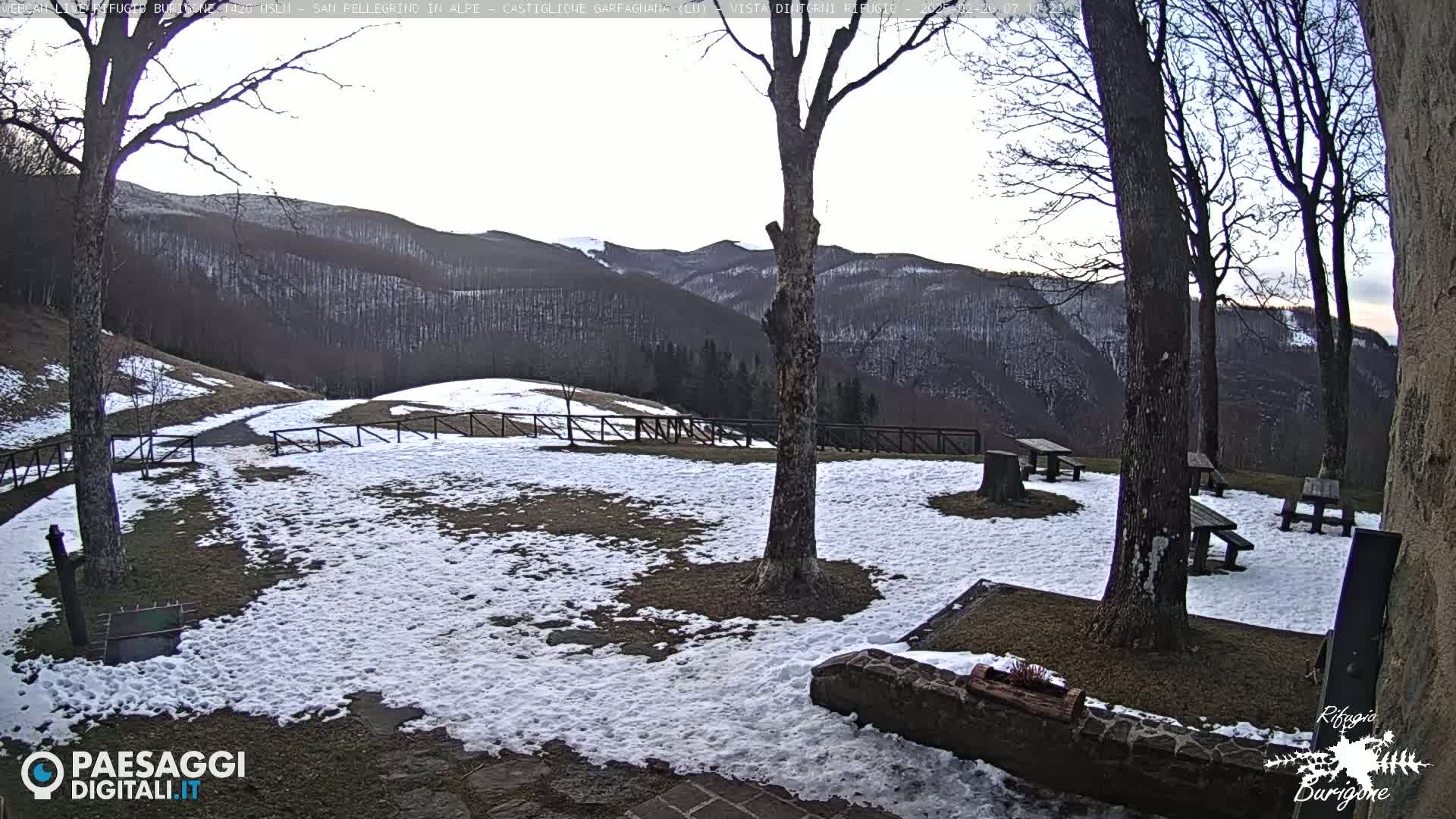 A snow-dusted outdoor area with several picnic tables sits beneath bare trees overlooking a mountain range under a pale sky.