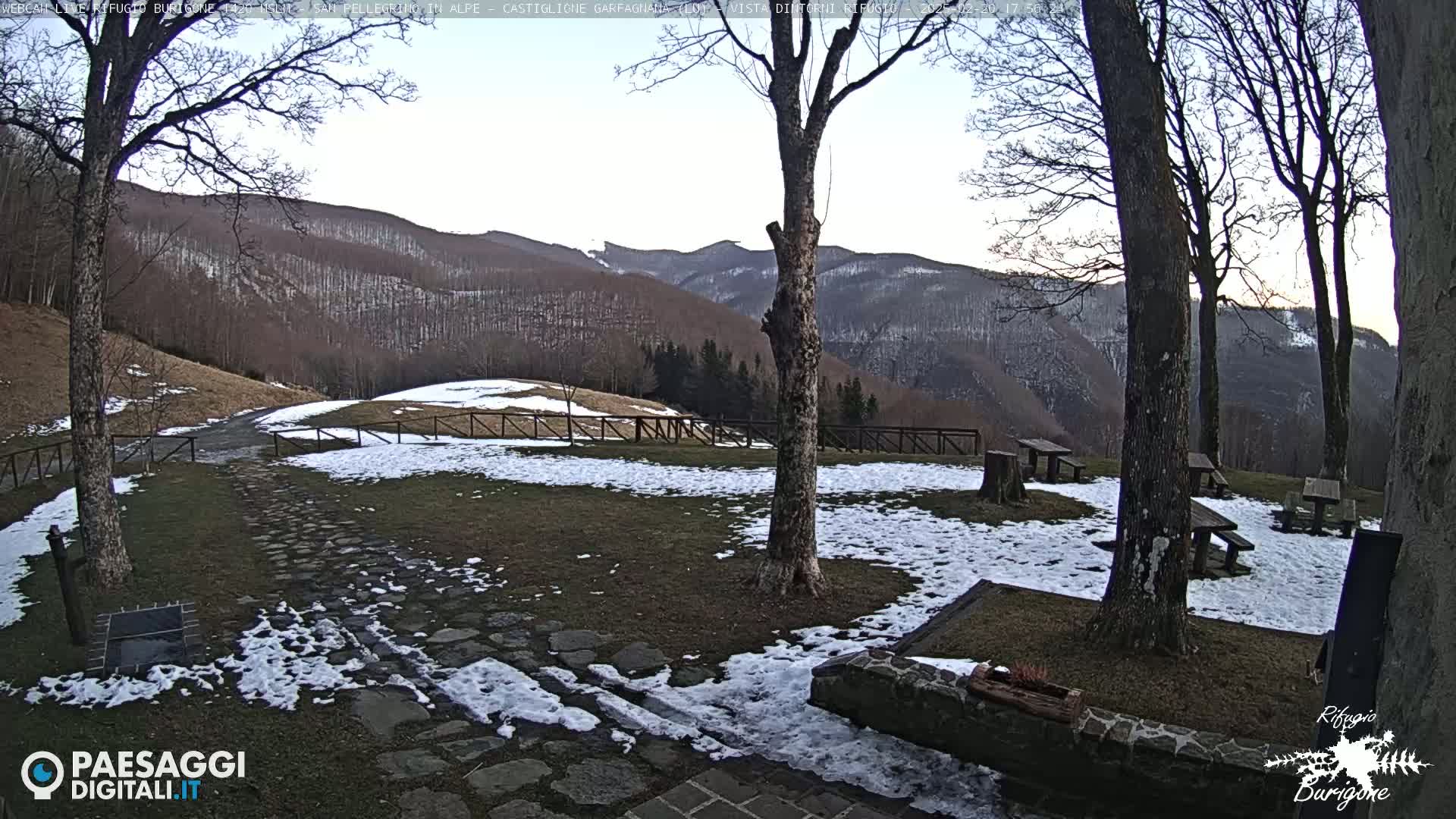 A snow-dusted grassy area with stone pathways, wooden picnic tables, and several bare trees overlooks a mountain range under a clear, pale sky.