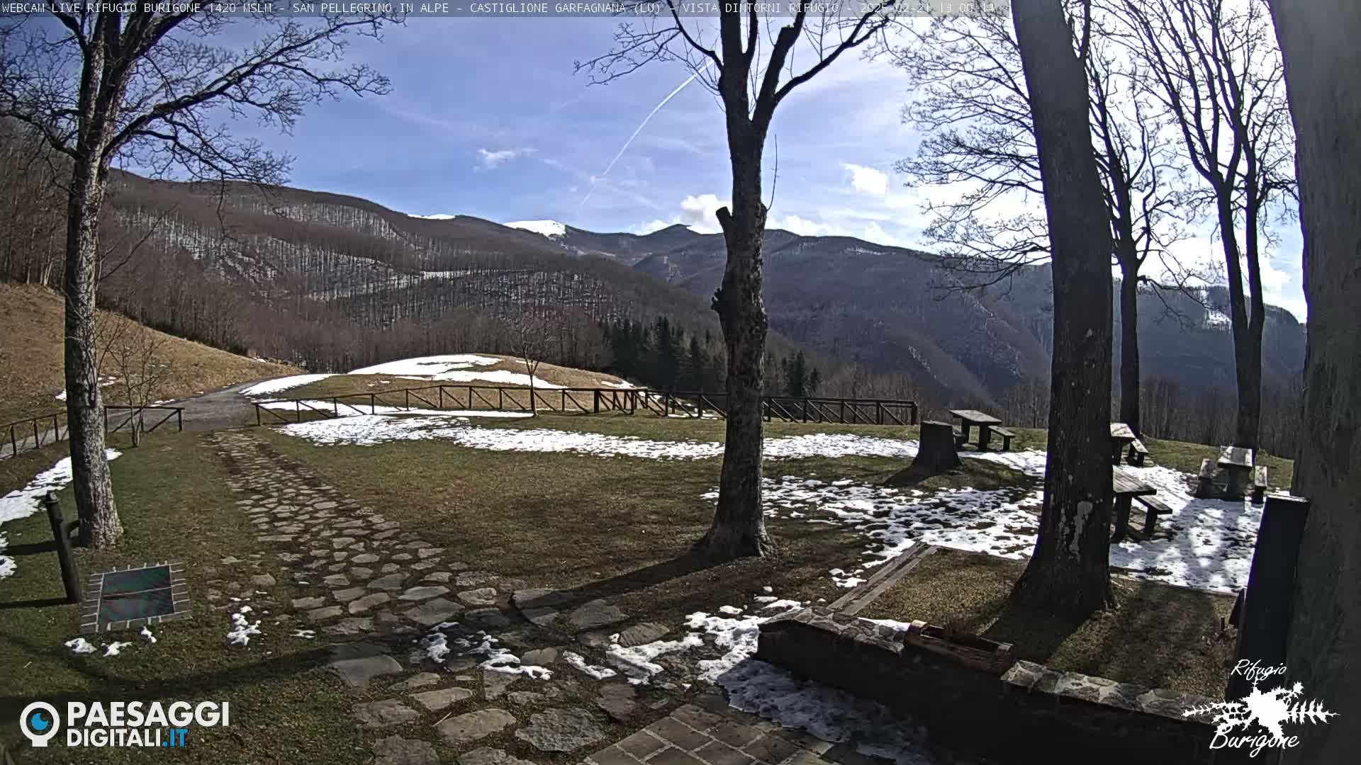 A stone path leads to picnic tables situated on a grassy area with patches of snow, overlooking a mountain range under a mostly sunny sky.