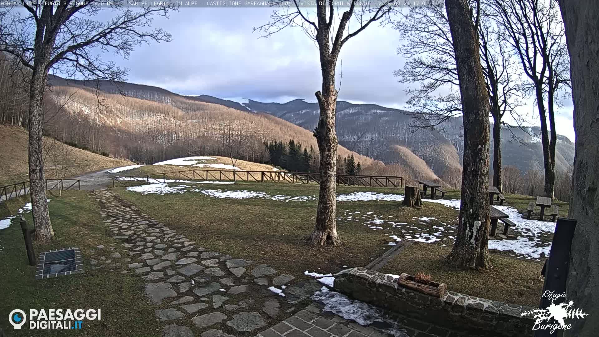 A stone path leads to picnic tables in a grassy area with snow patches, overlooking a mountain range under a partly cloudy sky.