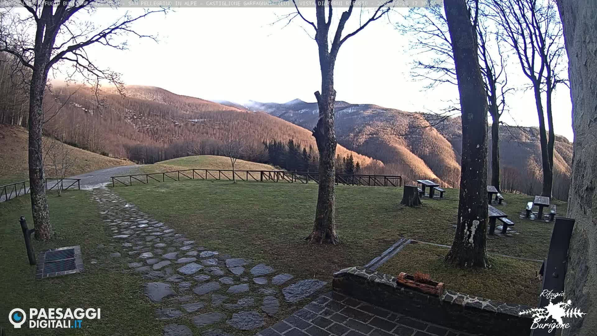 A stone path leads to picnic tables situated on a grassy area overlooking a range of snow-dusted mountains under a mostly clear sky.