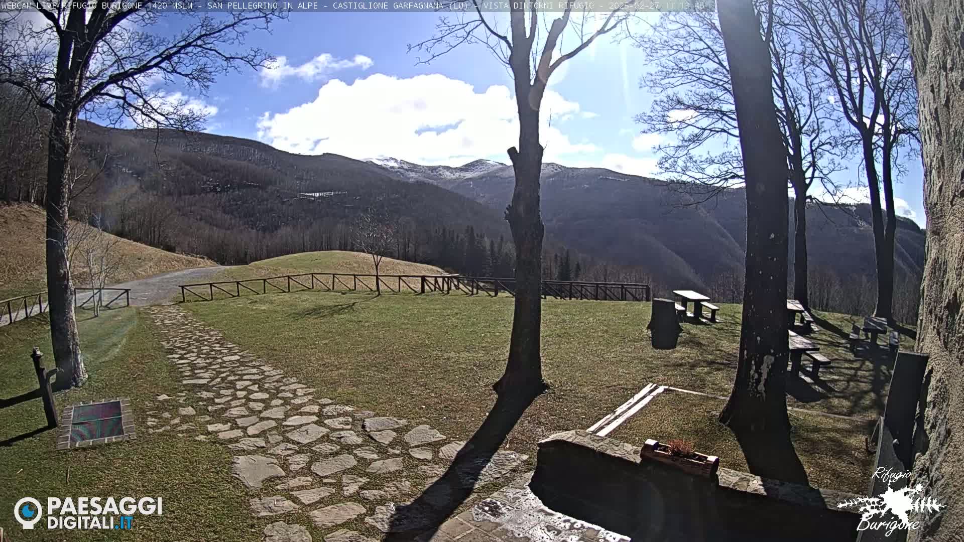 A stone path leads to a grassy area with picnic tables and trees, overlooking snow-capped mountains under a partly sunny sky.