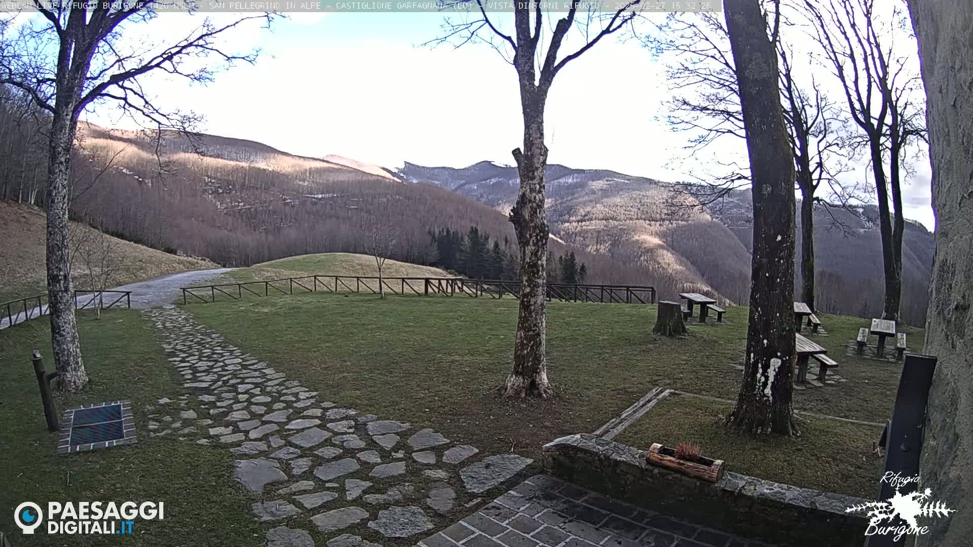 A stone path leads to several picnic tables situated on a grassy area overlooking a mountain range under a mostly sunny sky.