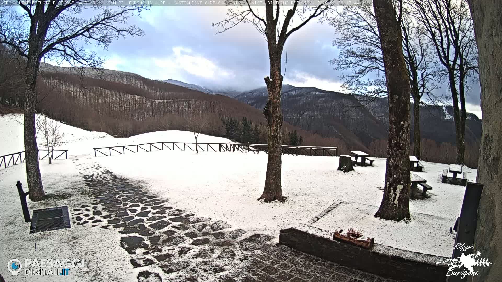 A snow-covered stone path leads to picnic tables beneath bare trees overlooking a snow-dusted mountain range under a cloudy sky.