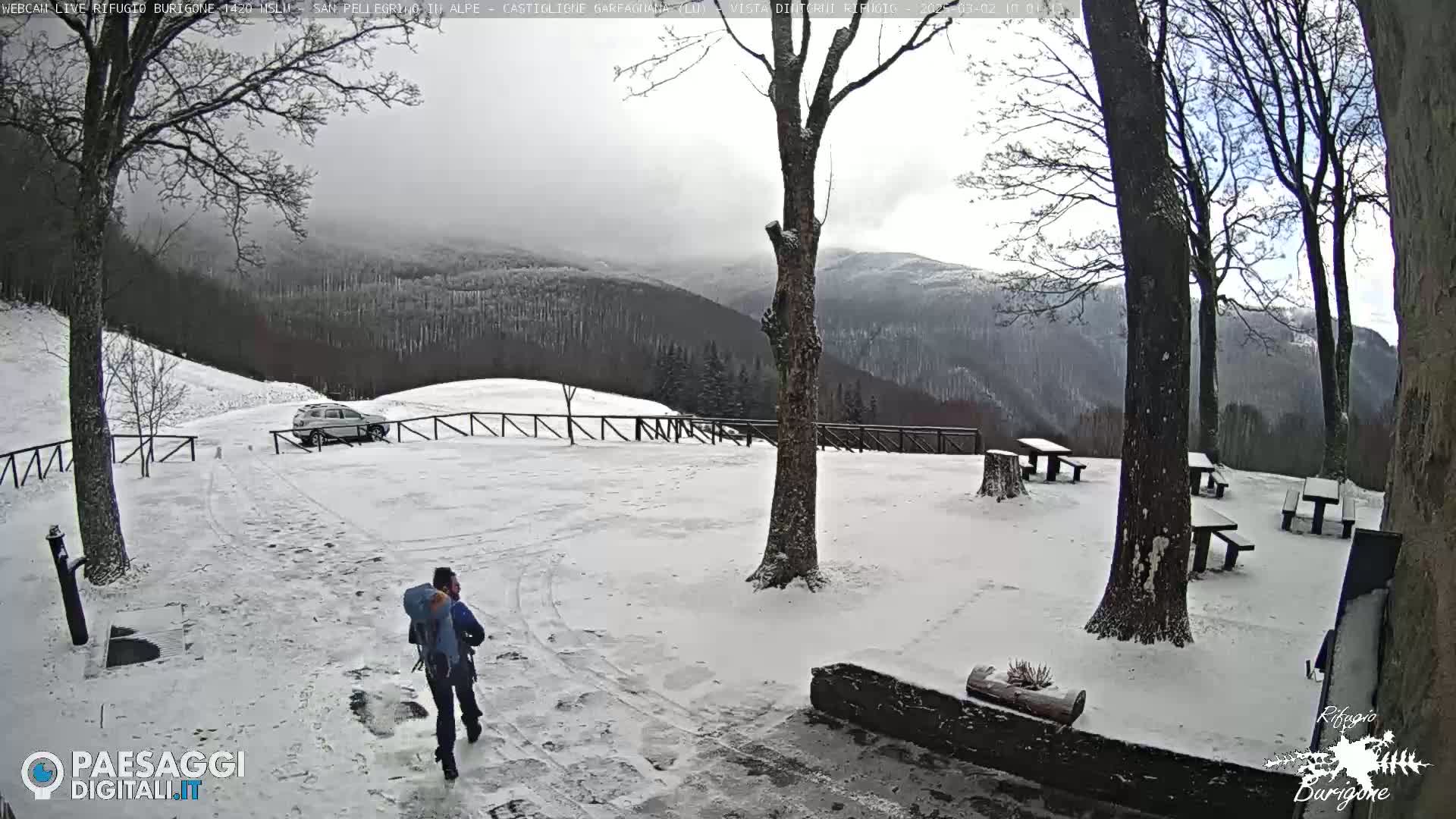 A person with a backpack walks on a snow-covered path toward a parking area with a vehicle, in front of a snowy mountain landscape with several picnic tables.