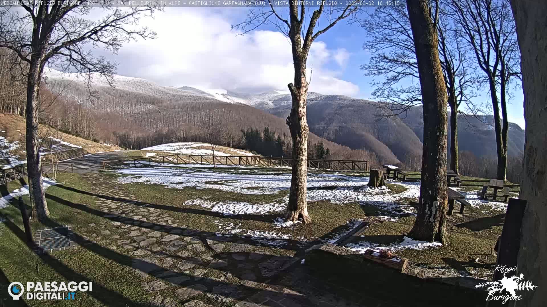 A partly sunny winter scene shows a snow-dusted grassy area with stone pathways, picnic tables, and trees, overlooking snow-capped mountains in the distance.