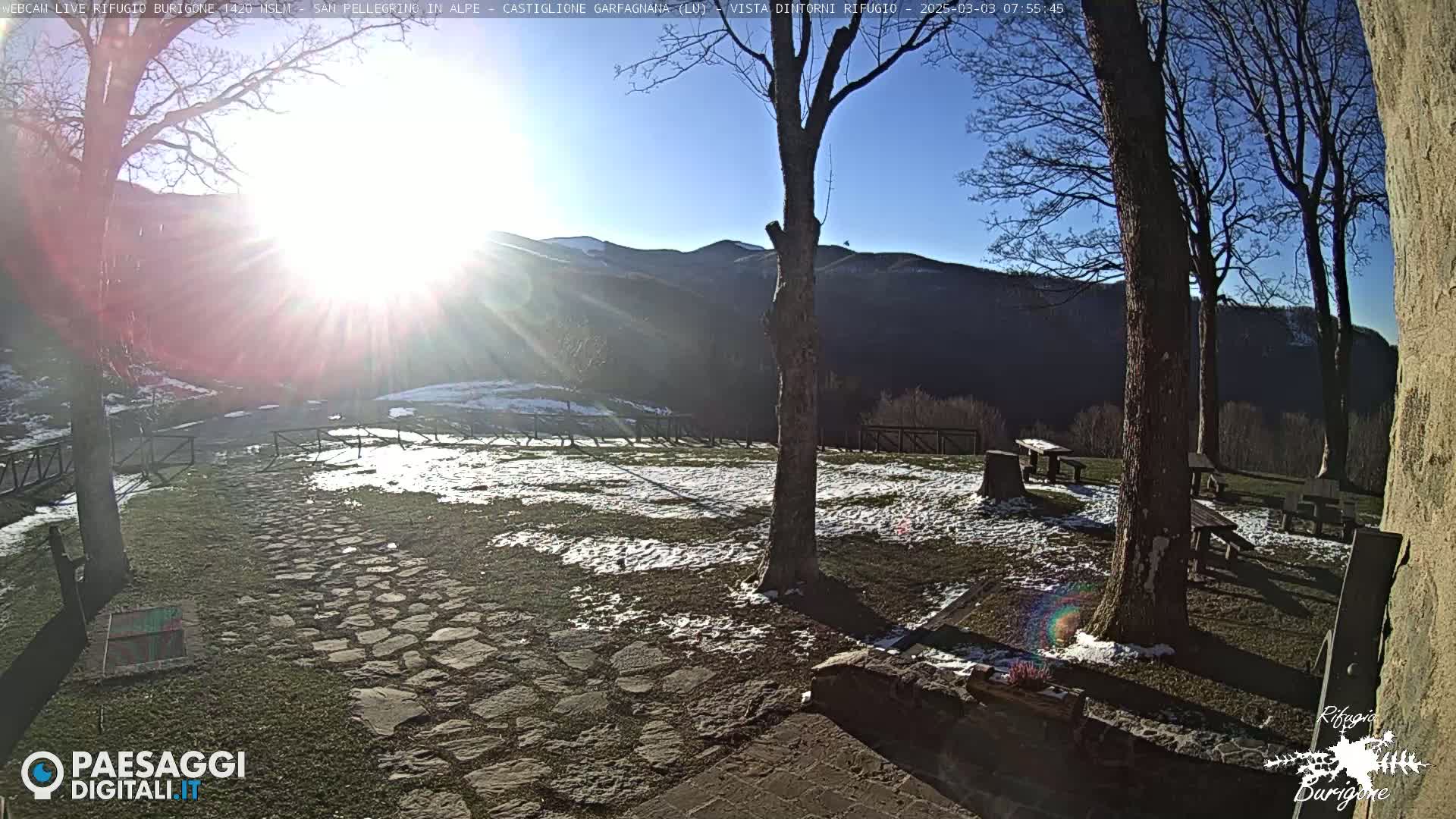 A sunny, cold day with patchy snow on the ground reveals a stone path leading to picnic tables situated in a grassy area near trees and mountains in the background.