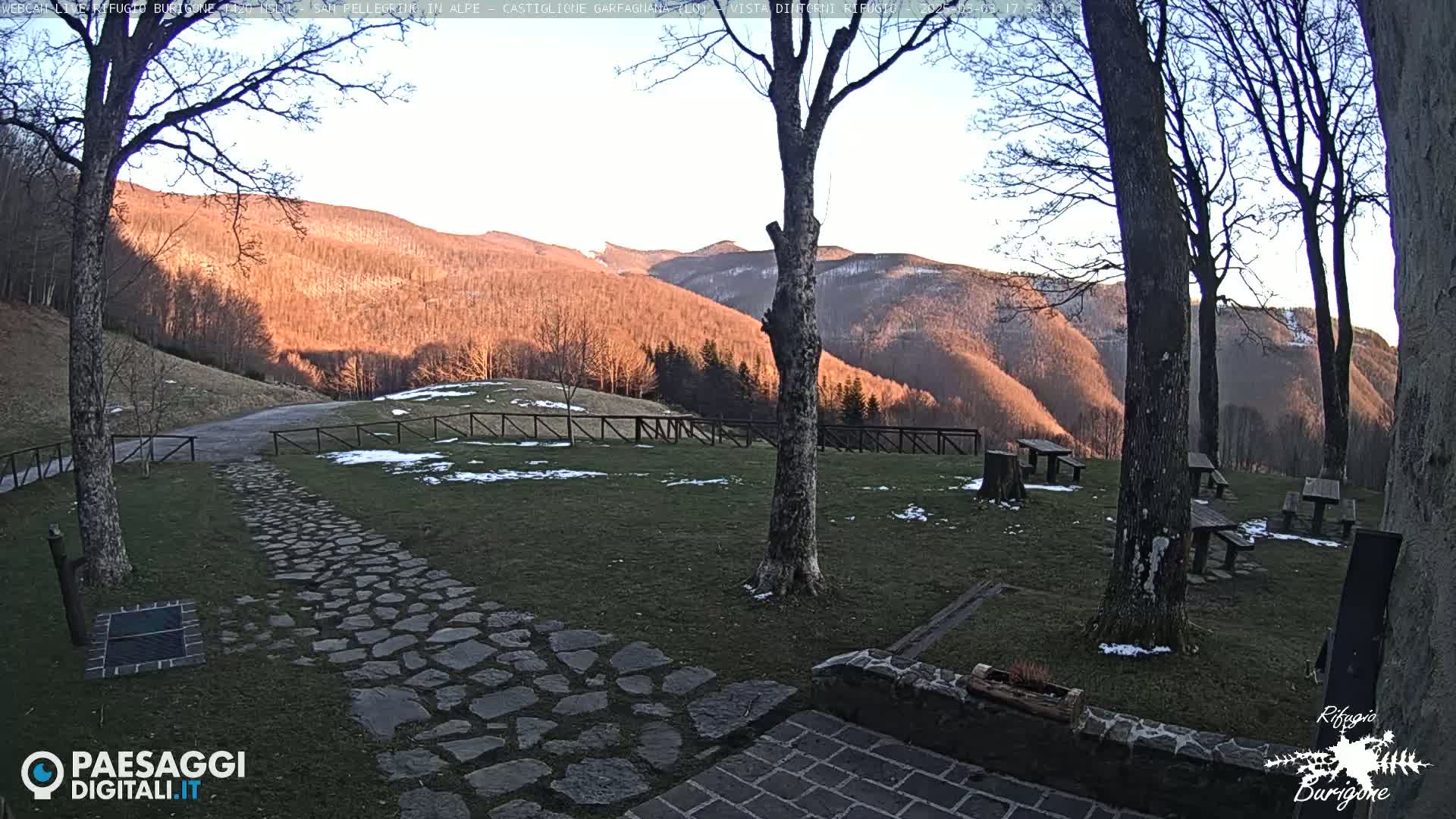 A stone path leads to a grassy area with picnic tables beneath bare trees, overlooking a sunlit hillside with patches of snow under a clear sky.