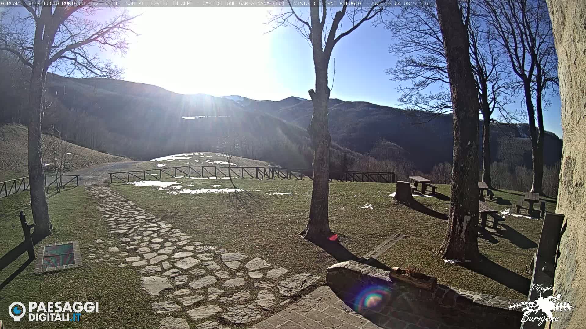 A stone path leads to wooden picnic tables situated on a grassy area with patches of snow, overlooking a mountain range under a clear, sunny sky.