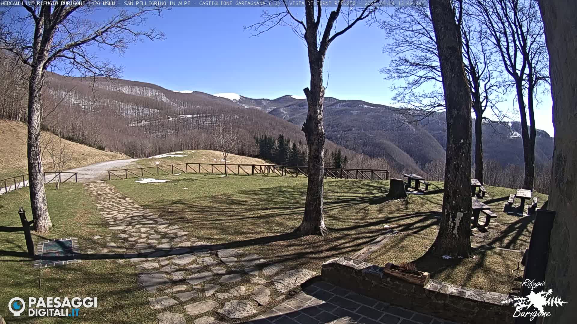 A stone path leads to picnic tables situated on a grassy area overlooking snow-dusted mountains under a clear blue sky.