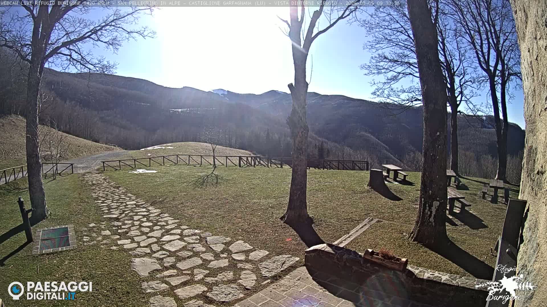 A sunny, stone pathway leads to a grassy area with picnic tables, wooden fences, and distant mountains with some snow visible under a clear blue sky.