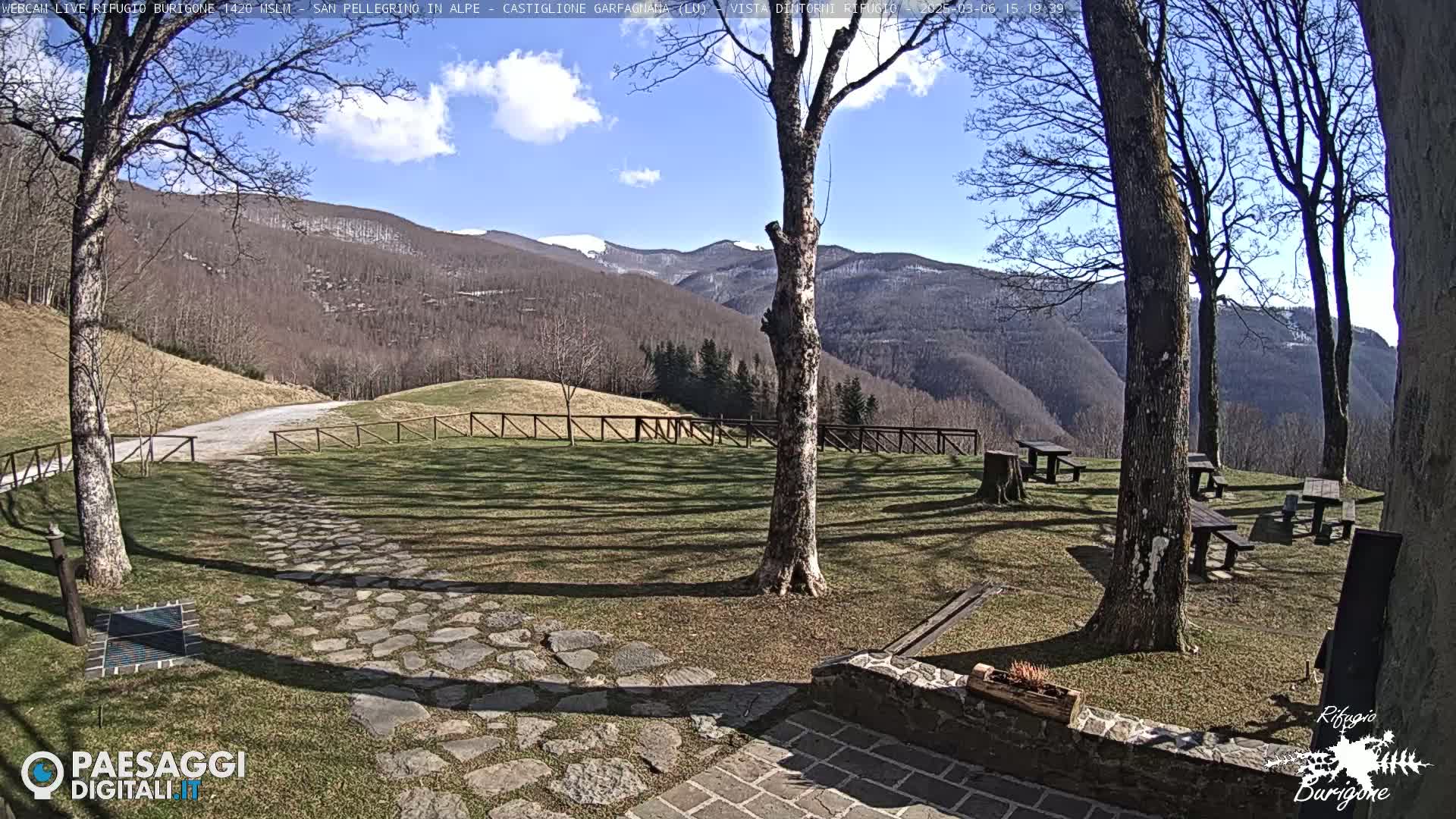 A sunny day reveals a stone pathway leading to picnic tables nestled in a grassy area, surrounded by leafless trees and overlooking snow-dusted mountains in the distance.
