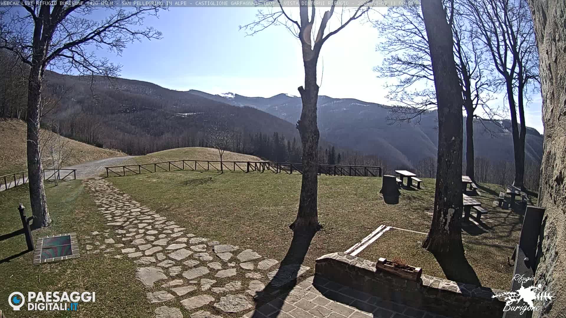 A stone path leads to a grassy area with several picnic tables situated beneath bare trees, overlooking a mountain range under a clear, sunny sky.
