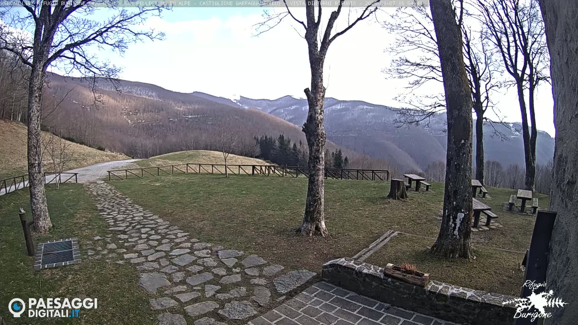 A stone path leads to picnic tables situated in a grassy area overlooking a mountain range under a mostly clear sky.