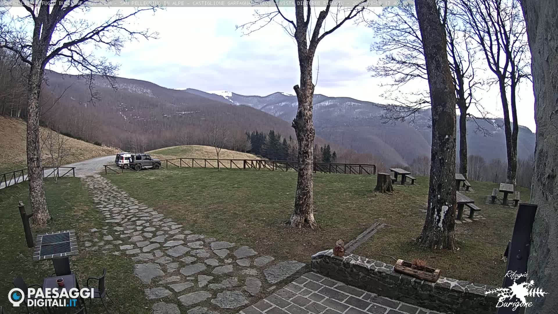 A stone path leads to a grassy area with picnic tables, a few cars, and trees, overlooking snow-capped mountains under a mostly cloudy sky.
