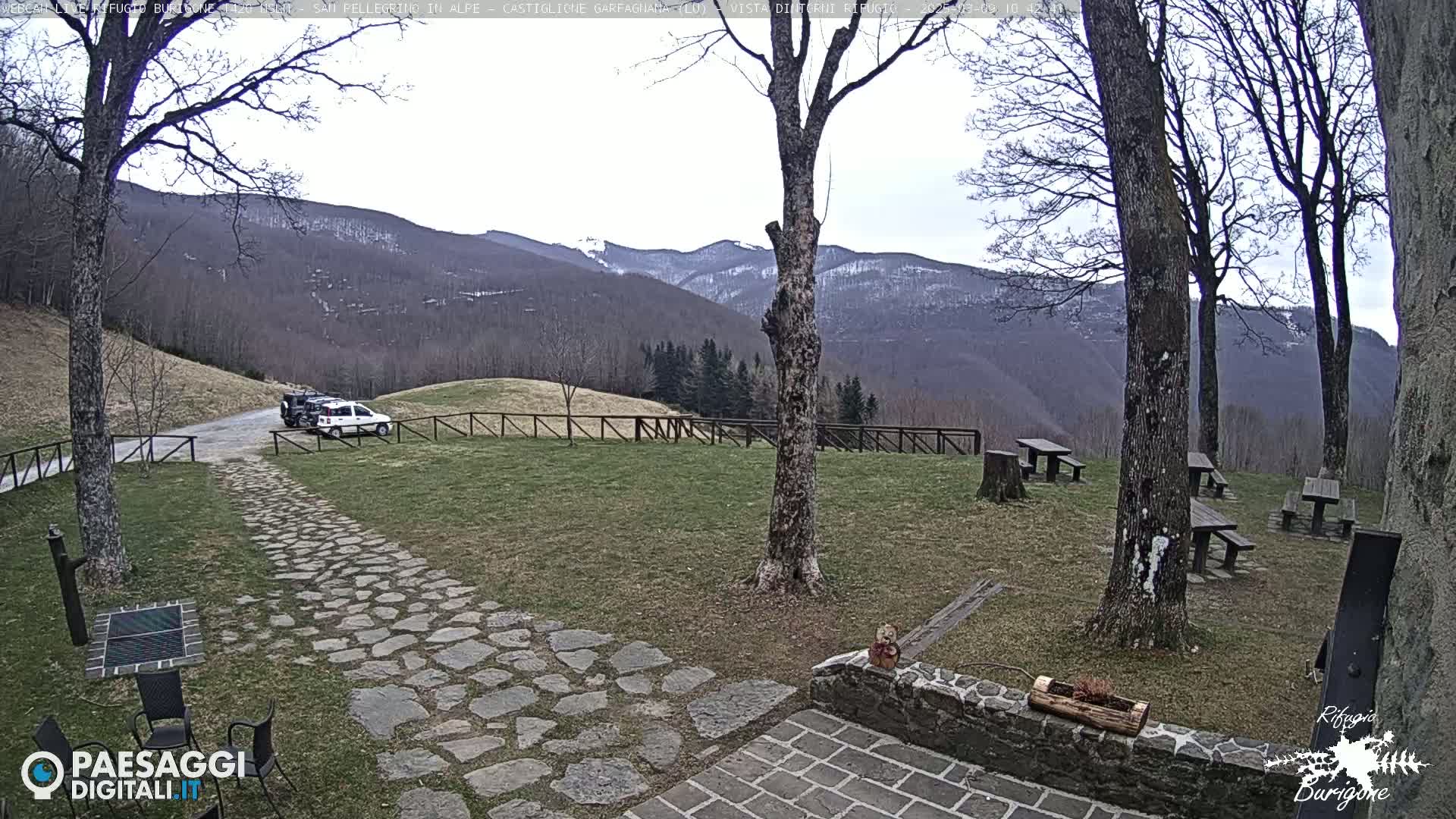 A stone path leads to a grassy area with picnic tables and a vehicle parked near a wooden fence, all set against a backdrop of distant mountains with some snow, under an overcast sky.
