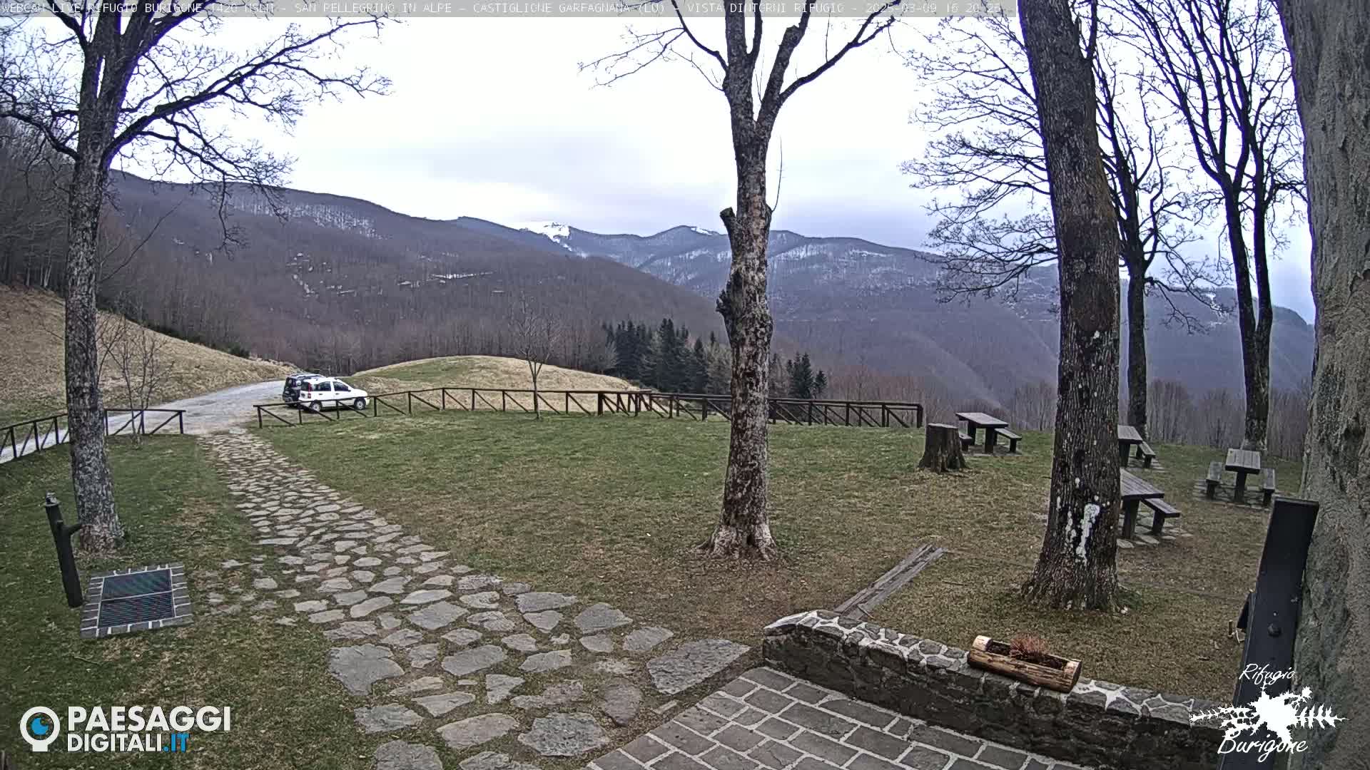 A stone path leads to a grassy area with picnic tables and a white car parked nearby, overlooking a mountain range with some snow under an overcast sky.