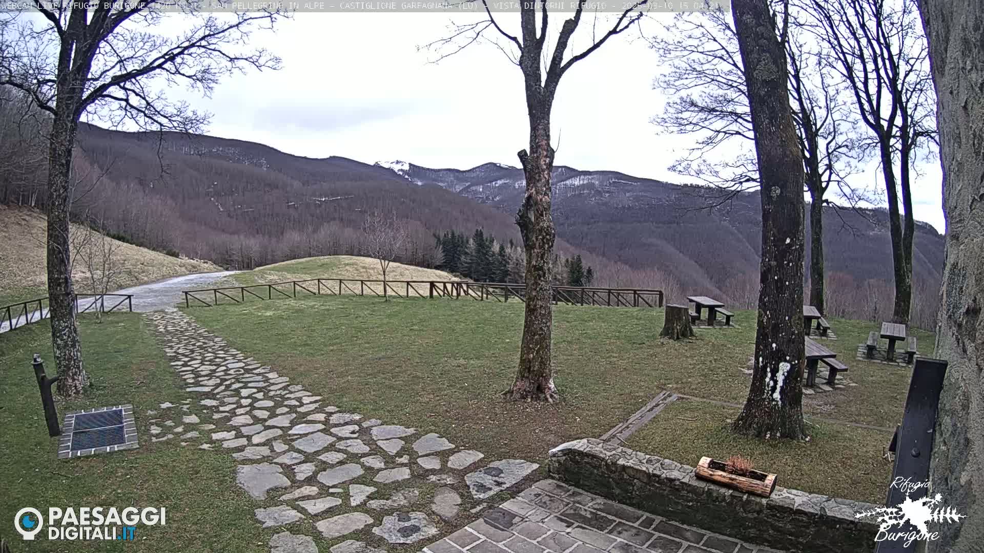 A stone path leads to picnic tables situated on a grassy area overlooking a mountain range with some snow, under a mostly cloudy sky.