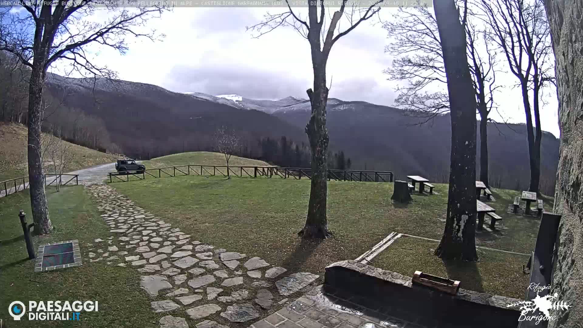 A stone path leads to a grassy area with picnic tables and a vehicle parked nearby, overlooking snow-capped mountains under a partly cloudy sky.