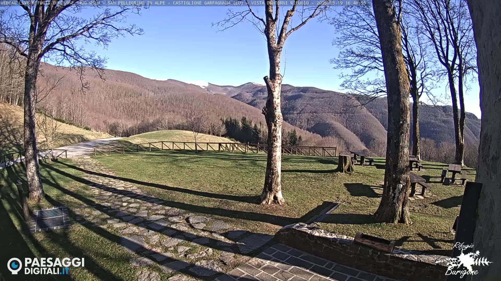 A sunny outdoor scene shows a grassy area with several picnic tables, a stone path, and several leafless trees, all set against a backdrop of a mountainous landscape with some remaining snow.