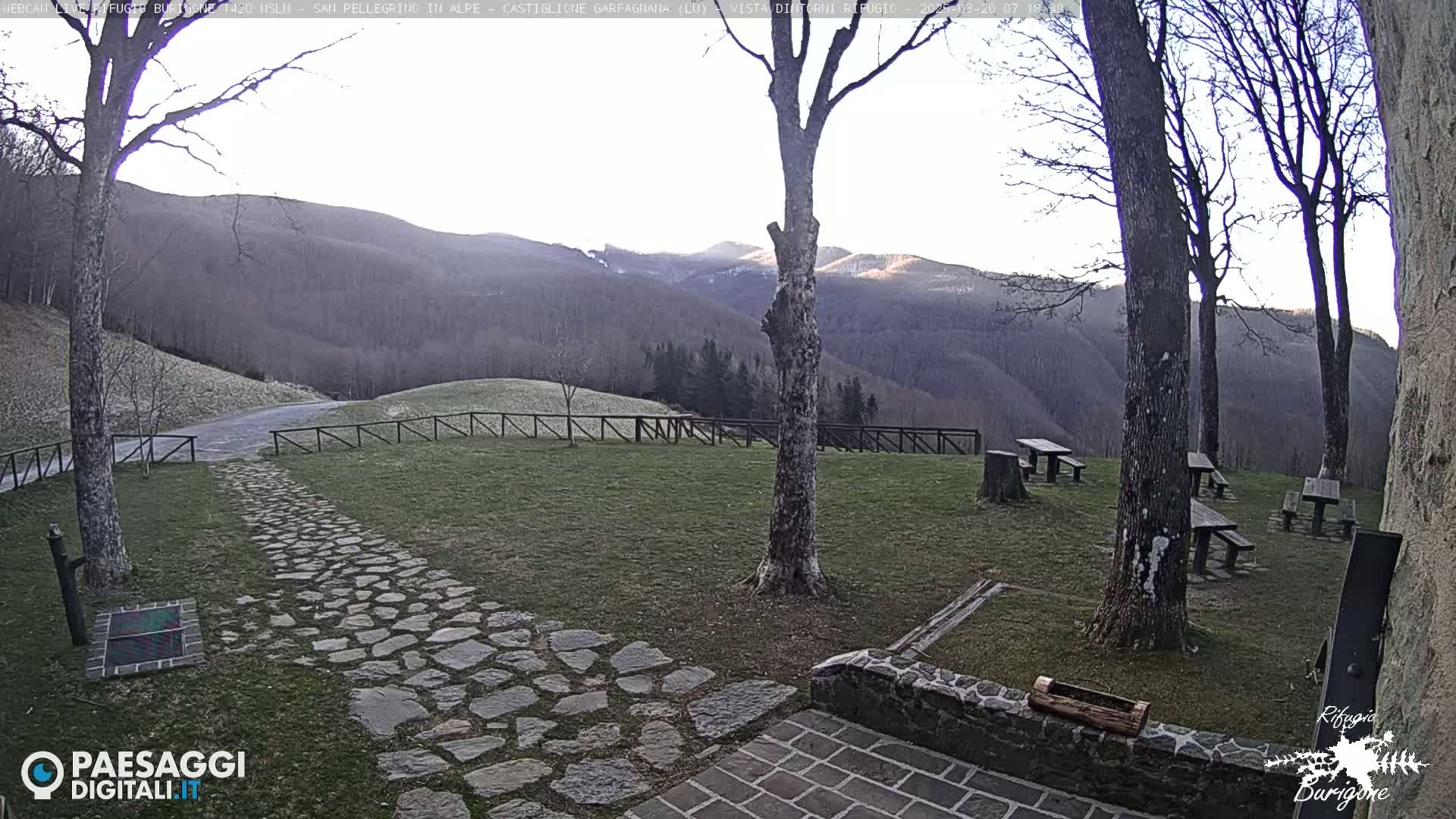 A stone path leads to a grassy area with picnic tables and trees, overlooking a mountain range under a mostly clear sky.