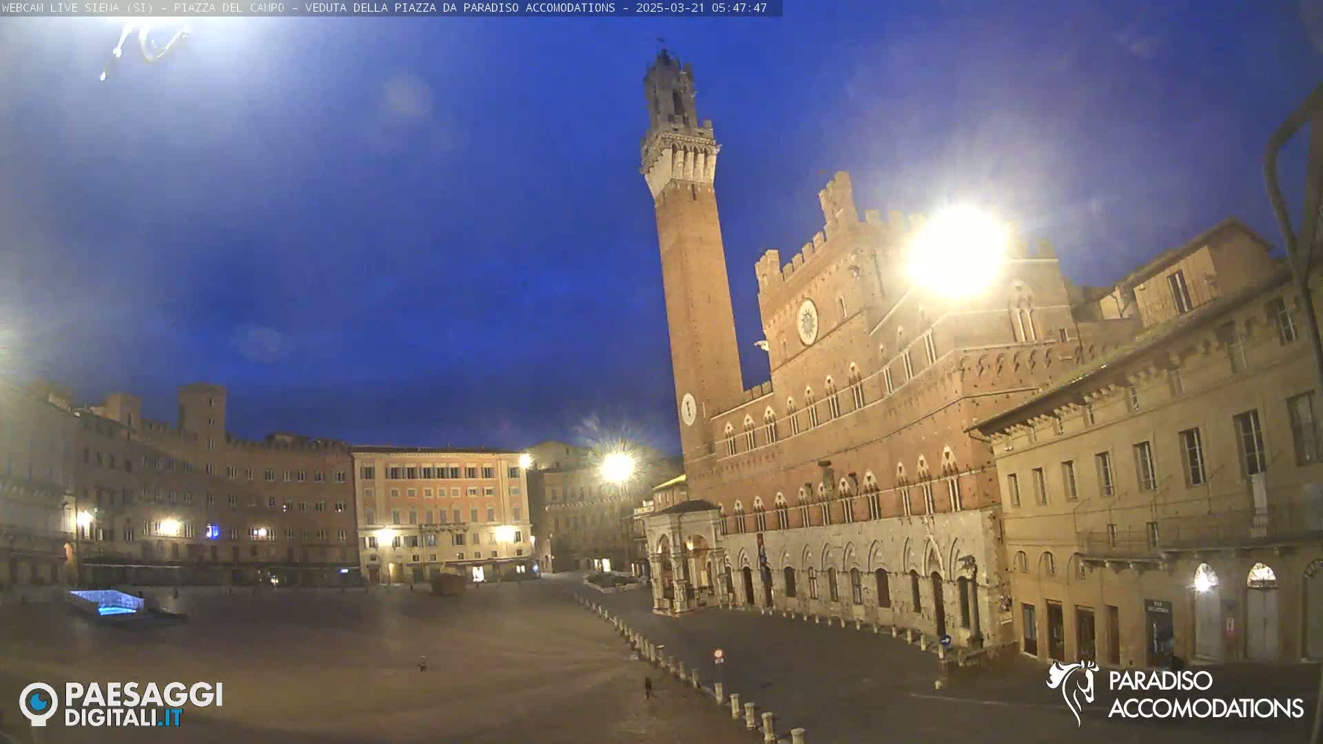 A twilight view of a large, mostly empty town square in Italy, dominated by a tall brick tower and adjacent medieval-style buildings under a clear, dark-blue sky.