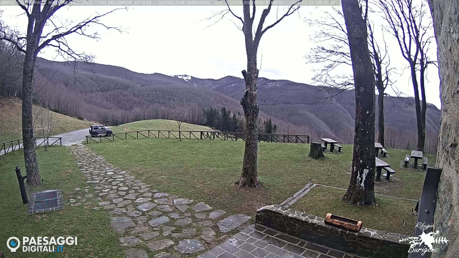 A stone pathway leads to a grassy area with picnic tables and a dark-colored SUV parked near a wooden fence, all overlooking a mountain range under a cloudy sky.