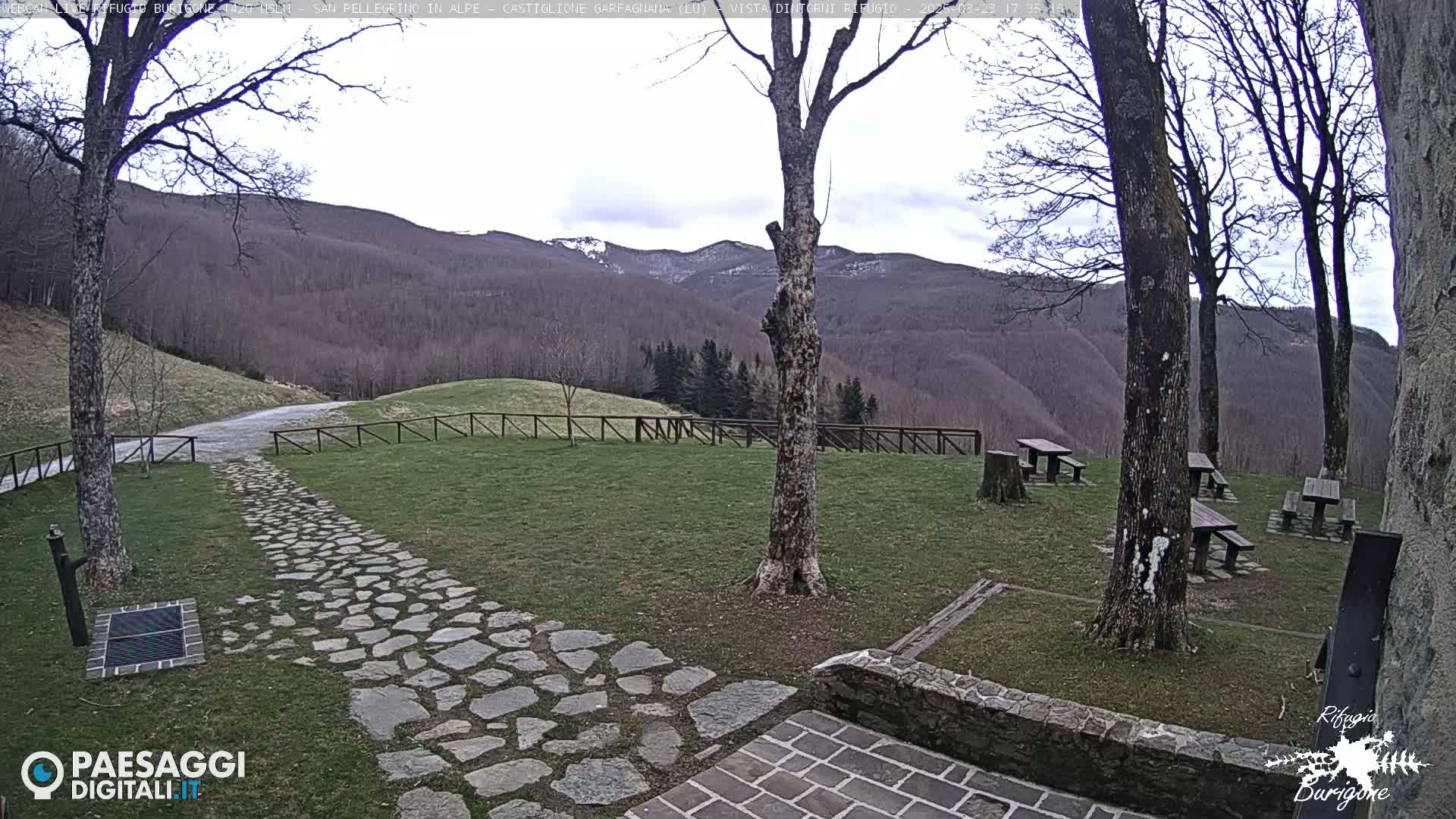 A stone path leads to several picnic tables situated on a grassy area overlooking a mountain range under a mostly cloudy sky.