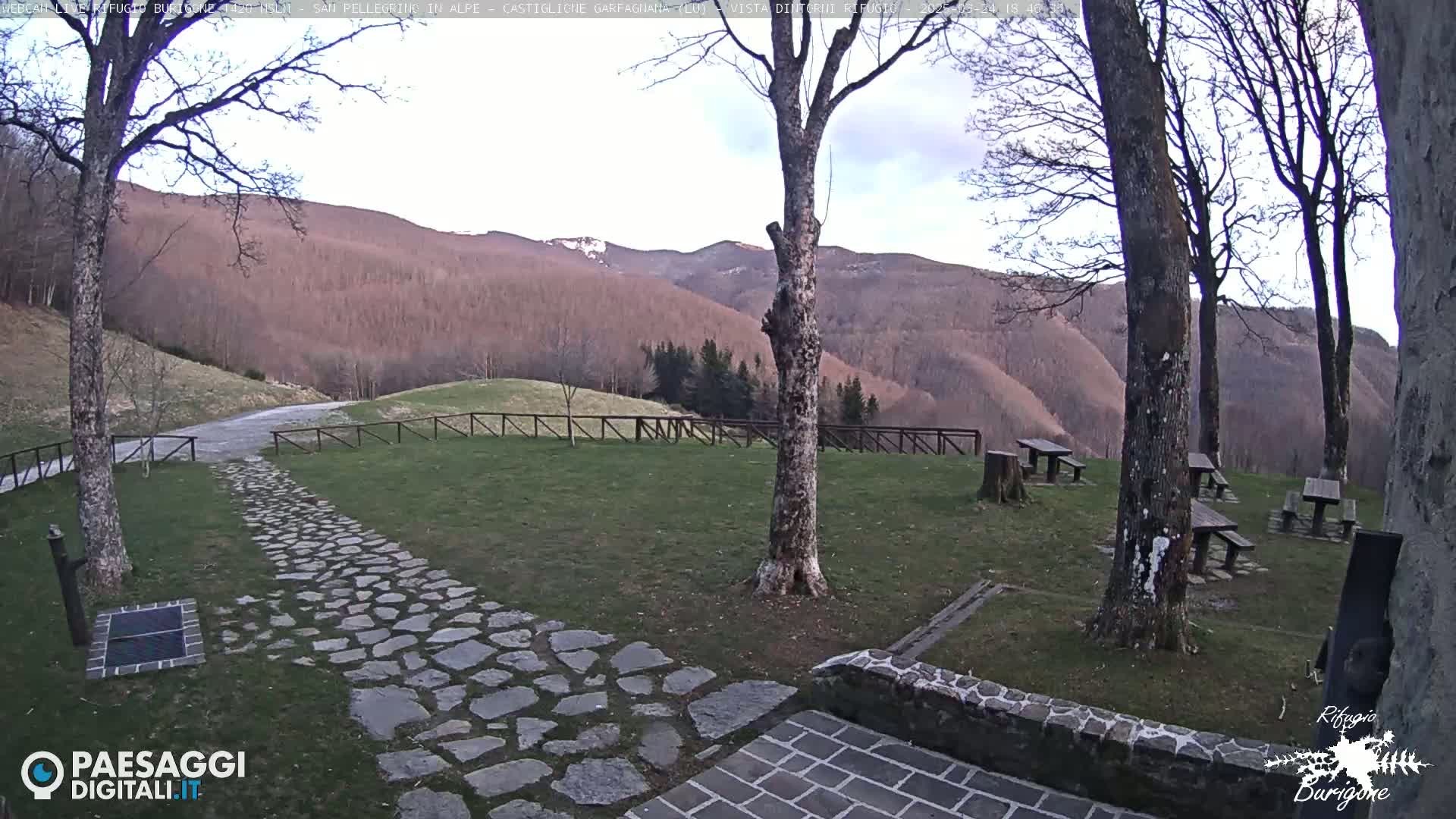 A stone pathway leads to picnic tables situated on a grassy area overlooking a mountain range under a partly cloudy sky.