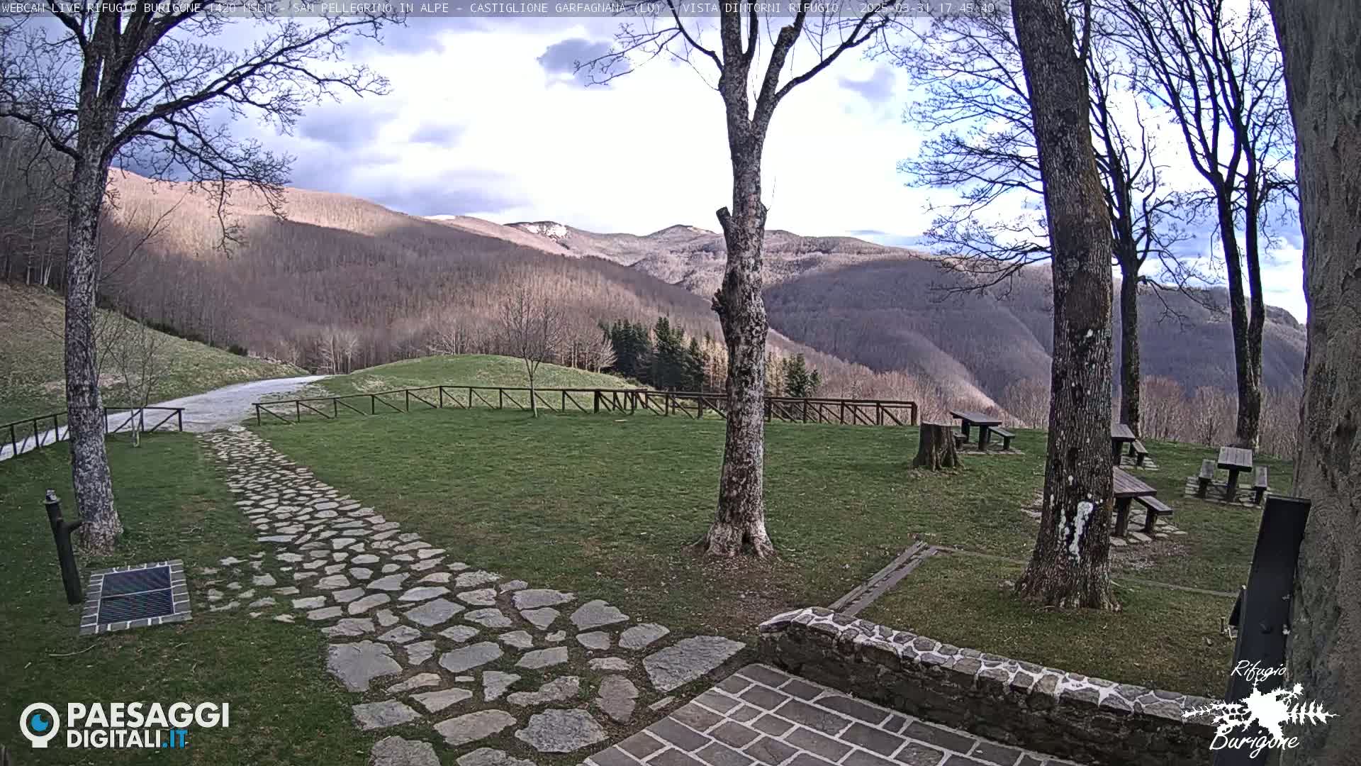 A stone path leads to a grassy area with picnic tables situated beneath leafless trees, overlooking a mountain range under a partly cloudy sky.