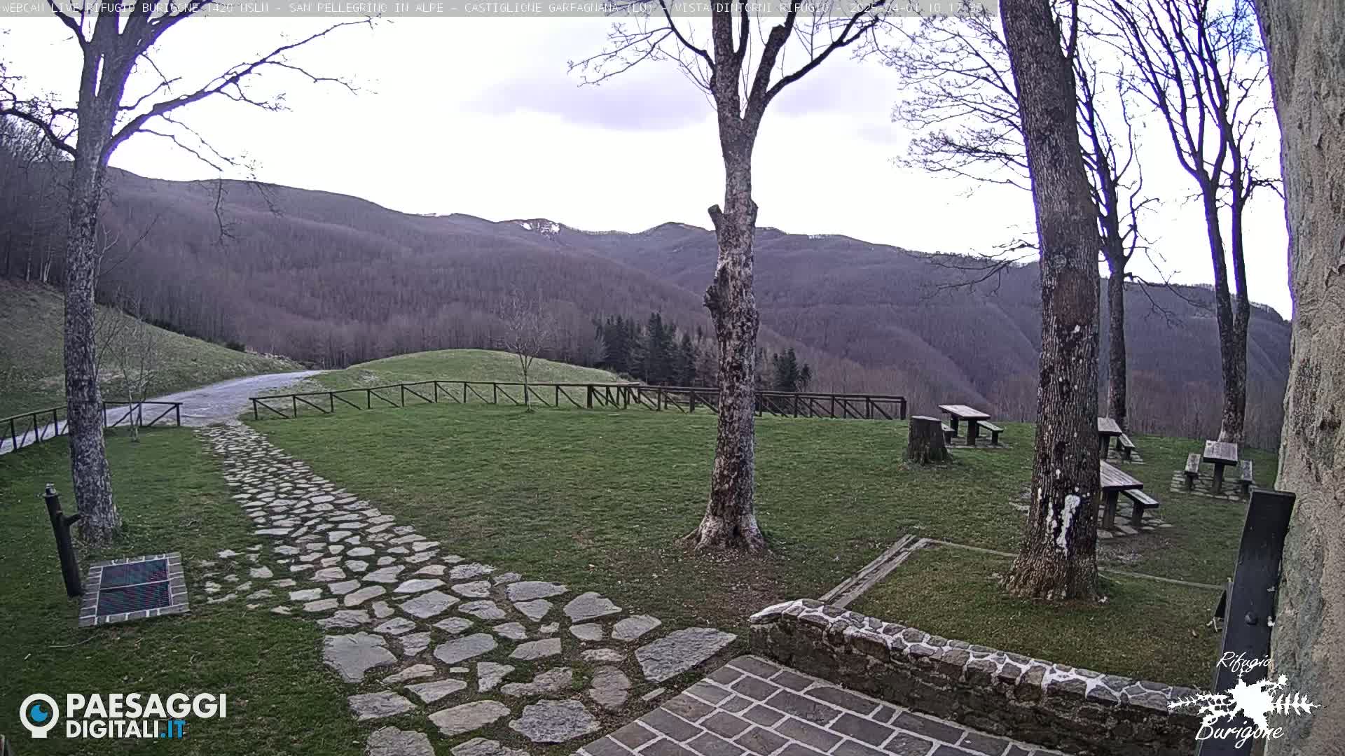 A stone path leads to picnic tables situated on a grassy area overlooking a mountain range under a partly cloudy sky.