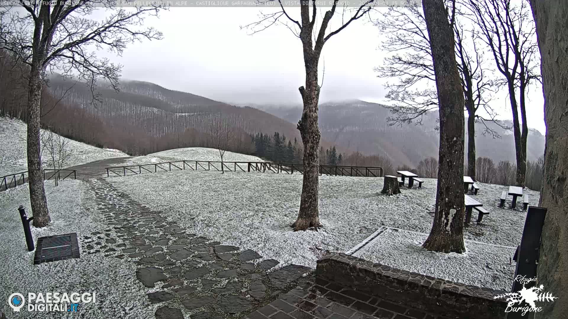 A snow-dusted stone pathway leads to several picnic tables situated in a clearing overlooking a mountain range under an overcast sky.
